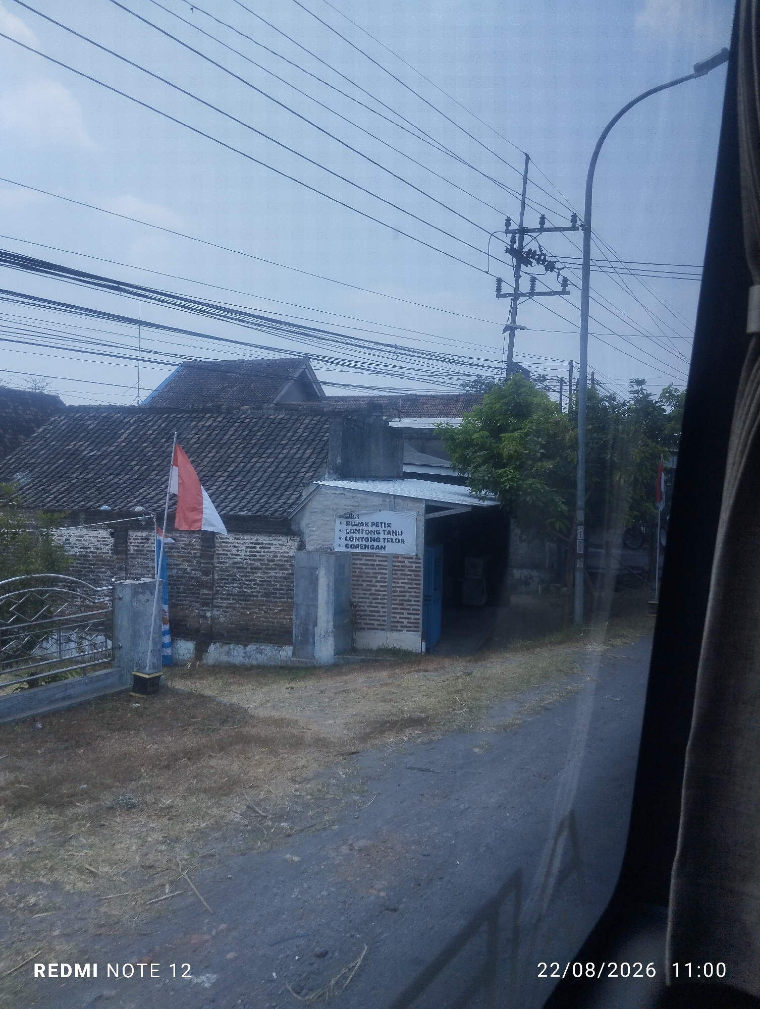 The image shows a street scene in Klagen Gambiran, Indonesia, on August 22, 2026, at 11:00 AM. The foreground is dominated by a rough asphalt road, with dry grass and scattered twigs on the side. In the mid-ground, there is a building with a tiled roof and brick walls. A small Indonesian flag is visible near the building. A sign on the building lists food items: "RUJAK PETIS", "LONTONG TAHU", "LONTONG SAYUR", and "GORENGAN". A few trees are also present. The background features a utility pole with numerous wires crisscrossing the sky. The overall lighting suggests an overcast day. The bottom left corner displays "REDMI NOTE 12".