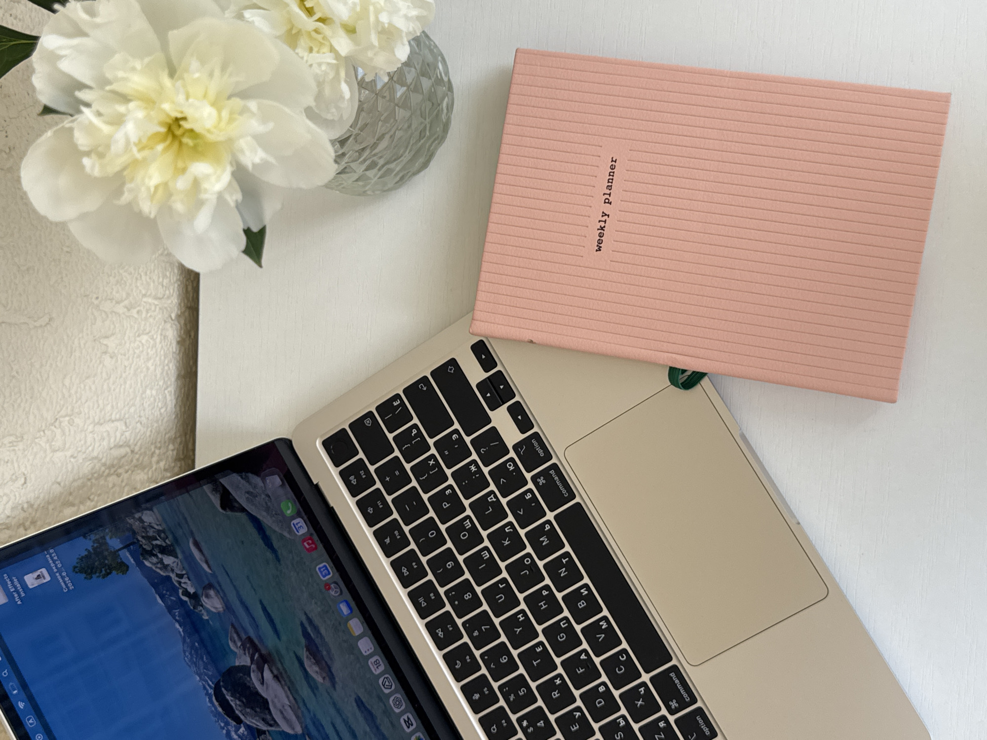 A top-down, eye-level shot shows a laptop and a pink notebook with the words "weekly planner" embossed on its cover, placed on a white wooden desk. The laptop is open, displaying a scenic wallpaper of a lake and mountains, with several app icons visible in the dock. A vase containing a bouquet of white peonies sits to the right of the laptop. The background features a textured white wall. The scene is devoid of any human presence or activity. The location is not identifiable as Yekaterinburg, Russia.