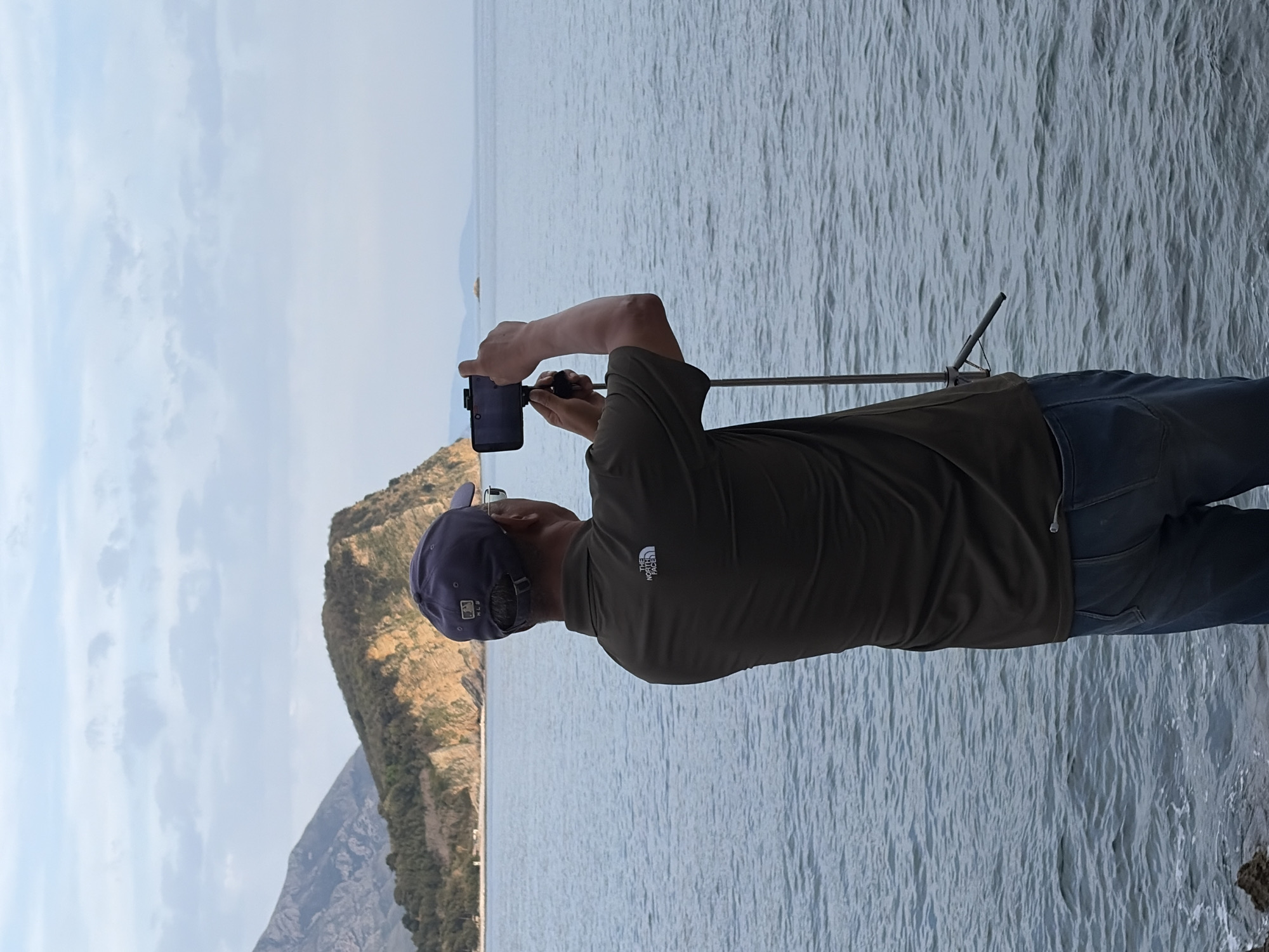 A man stands on the shore of the Adriatic Sea in Budva, Montenegro, using his smartphone on a tripod to capture the scene. He is wearing a dark green t-shirt with The North Face logo and blue jeans. He is also wearing a purple baseball cap. The sea is calm with small waves. In the background is a rocky peninsula. The sky is mostly clear with scattered clouds.
