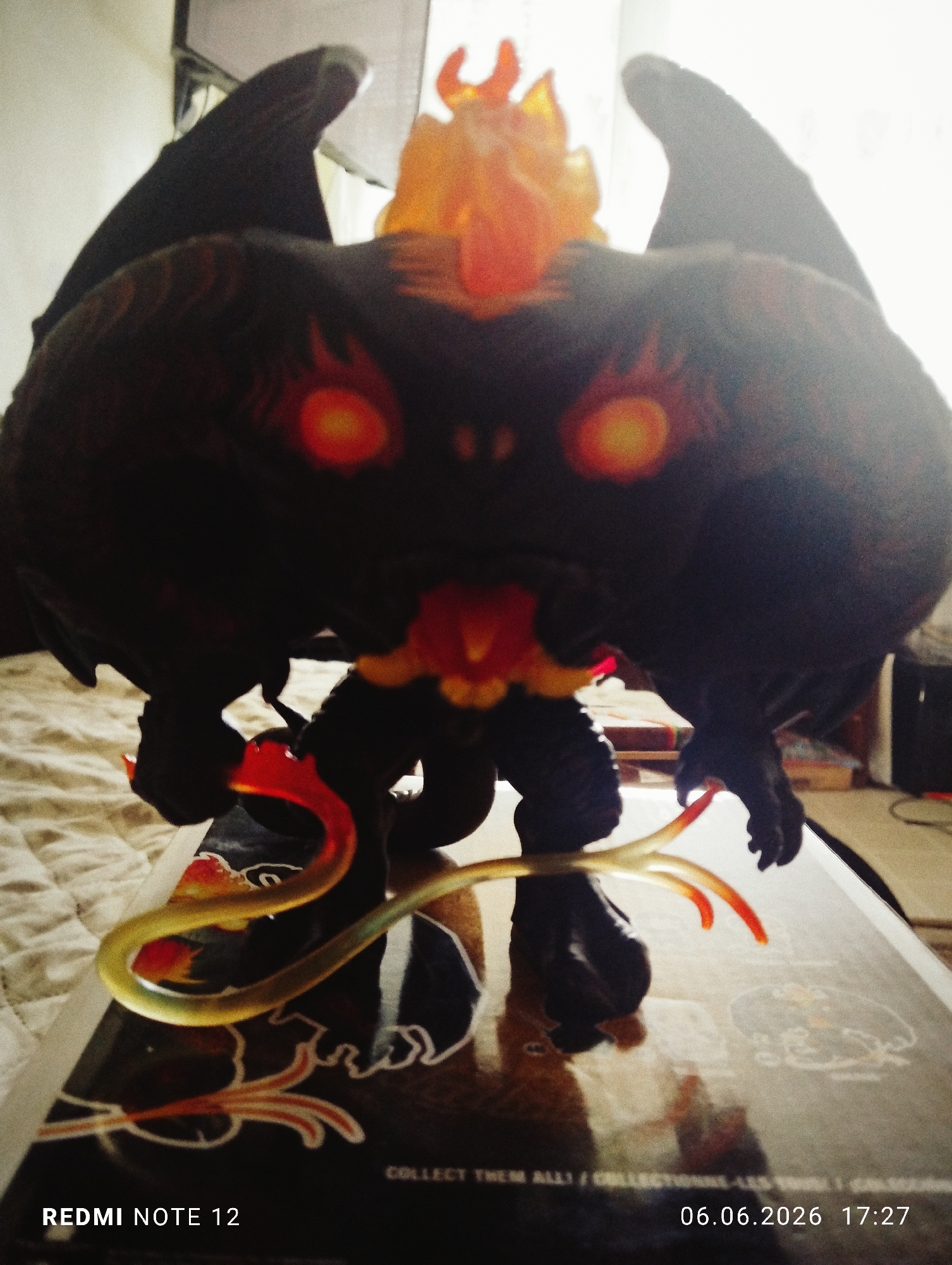A low-angle, backlit photograph depicts a stylized dark-colored figurine, resembling a Balrog from *The Lord of the Rings*, positioned on a reflective surface. The figurine features prominent wings, glowing orange eyes, and an orange-red fiery crown-like element on its head. A whip-like appendage, primarily yellow-orange with red tips, extends from its right hand. The figurine’s mouth area also exhibits a red glow. The reflective surface beneath the figurine displays text that includes "COLLECT THEM ALL!" and fragmented stylized fiery patterns, indicating it is likely product packaging or a display base. The background on the left consists of a light-colored, textured fabric, potentially a bedspread. The central and right background is intensely overexposed by a bright light source, likely a window, creating a strong silhouette effect around the figurine. No individuals are visible. The image was captured with a "REDMI NOTE 12" device on June 6, 2026, at 17:27. This scene is located in the city of Burgas, Bulgaria.