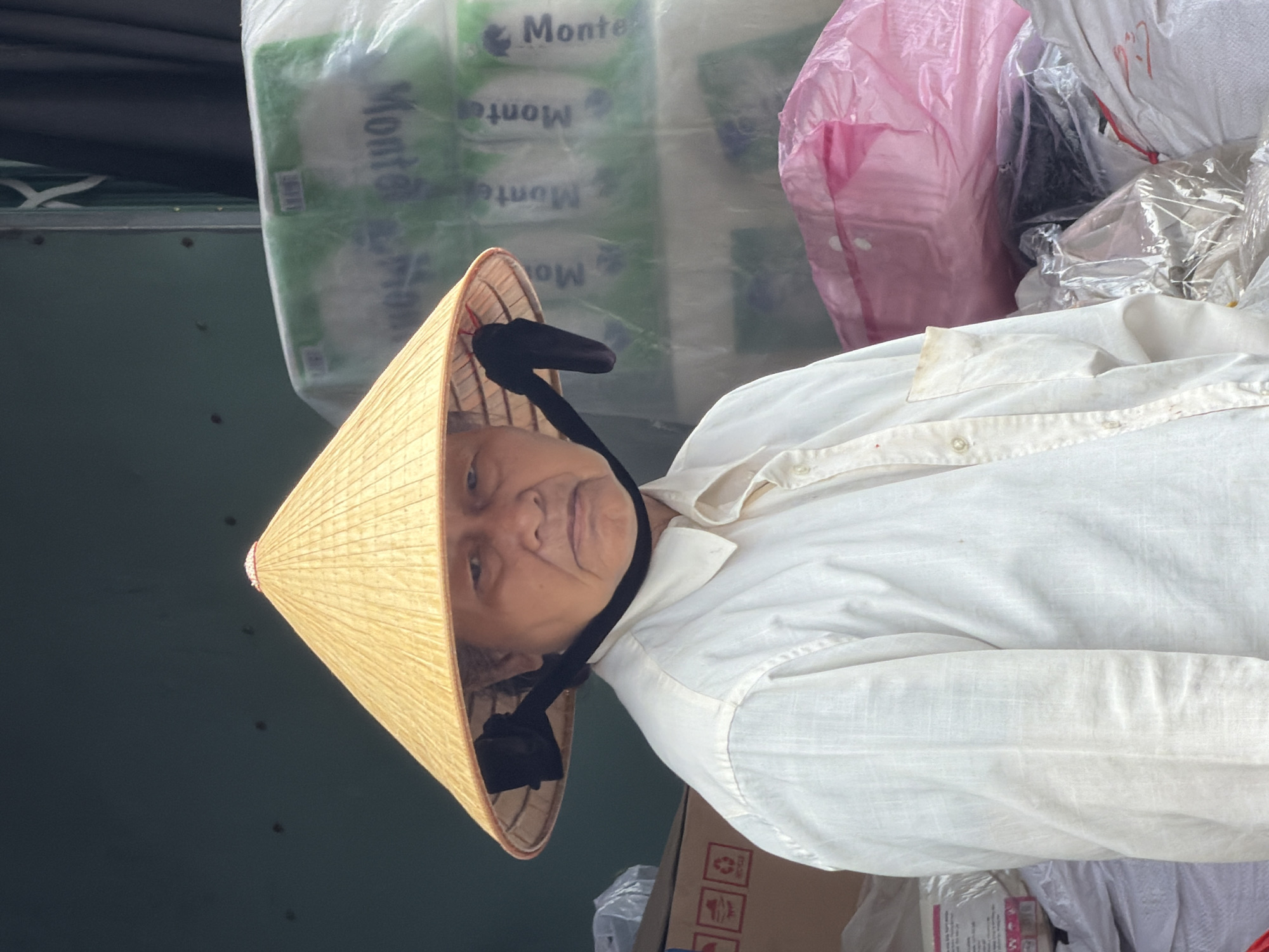 A person wearing a conical hat, a white linen shirt, and a dark strap across their chin stands outdoors. The hat is made of woven straw and is a light yellow color. The shirt is buttoned up to the collar and appears slightly wrinkled. Behind the person, stacked items are visible, including what appear to be large rolls of material wrapped in clear plastic, some with green and white labels that read "Monte". To the right of these rolls, a large pink plastic bag is visible. Further to the left, there is a cardboard box with red symbols. The ground is not visible. The overall scene suggests an outdoor market or storage area in Nha Trang, Vietnam.