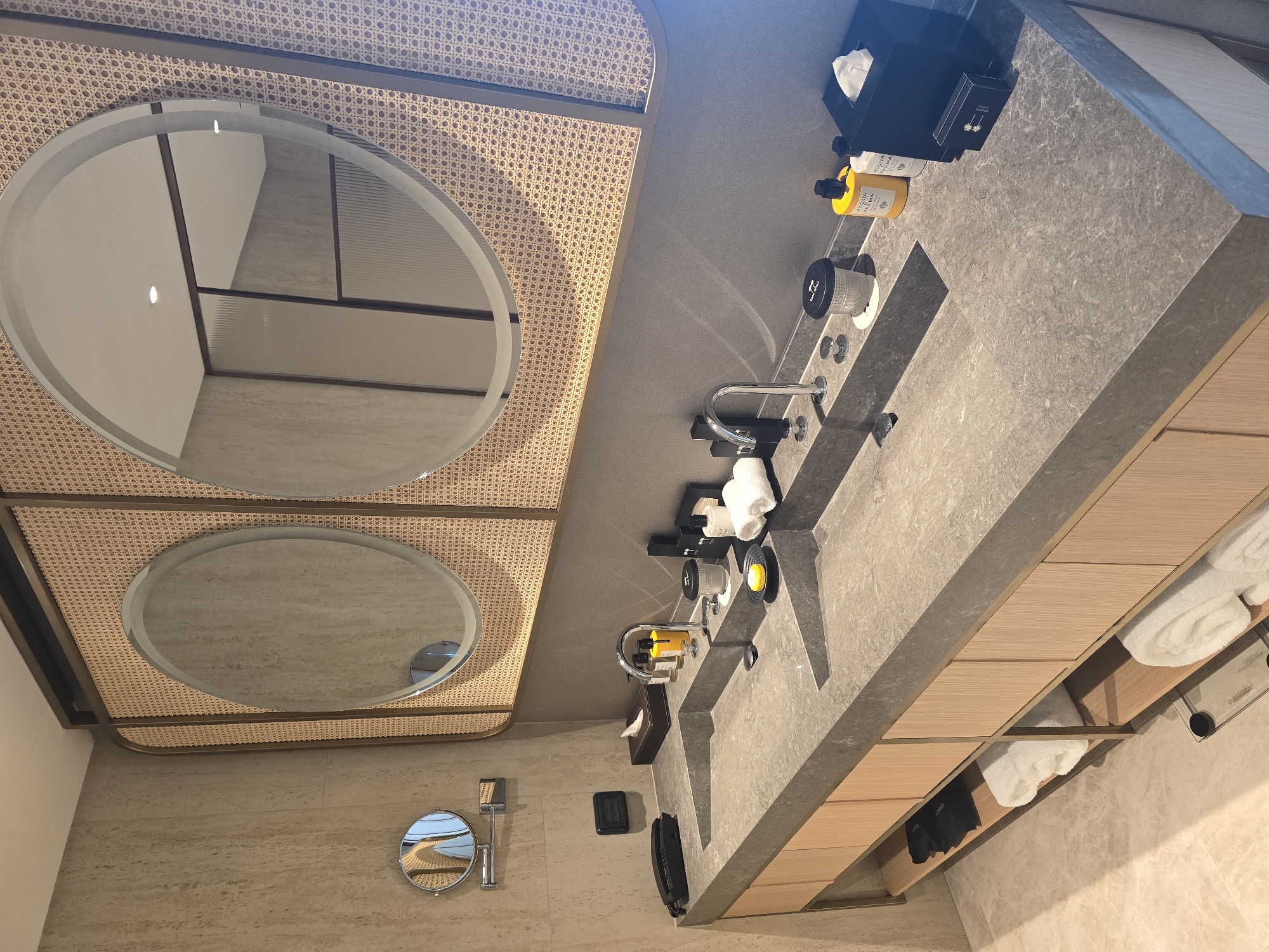The image shows a bathroom interior. Two oval mirrors are visible, framed by a woven design. A smaller, circular mirror is also present. A gray countertop features a sink and various bathroom amenities such as soap dispensers, towels, and toiletries. Wooden cabinetry is installed below the countertop. The walls are clad in a light-colored material. The scene depicts a bathroom setting, but lacks contextual information to confirm its location in Tel-Aviv, Israel.