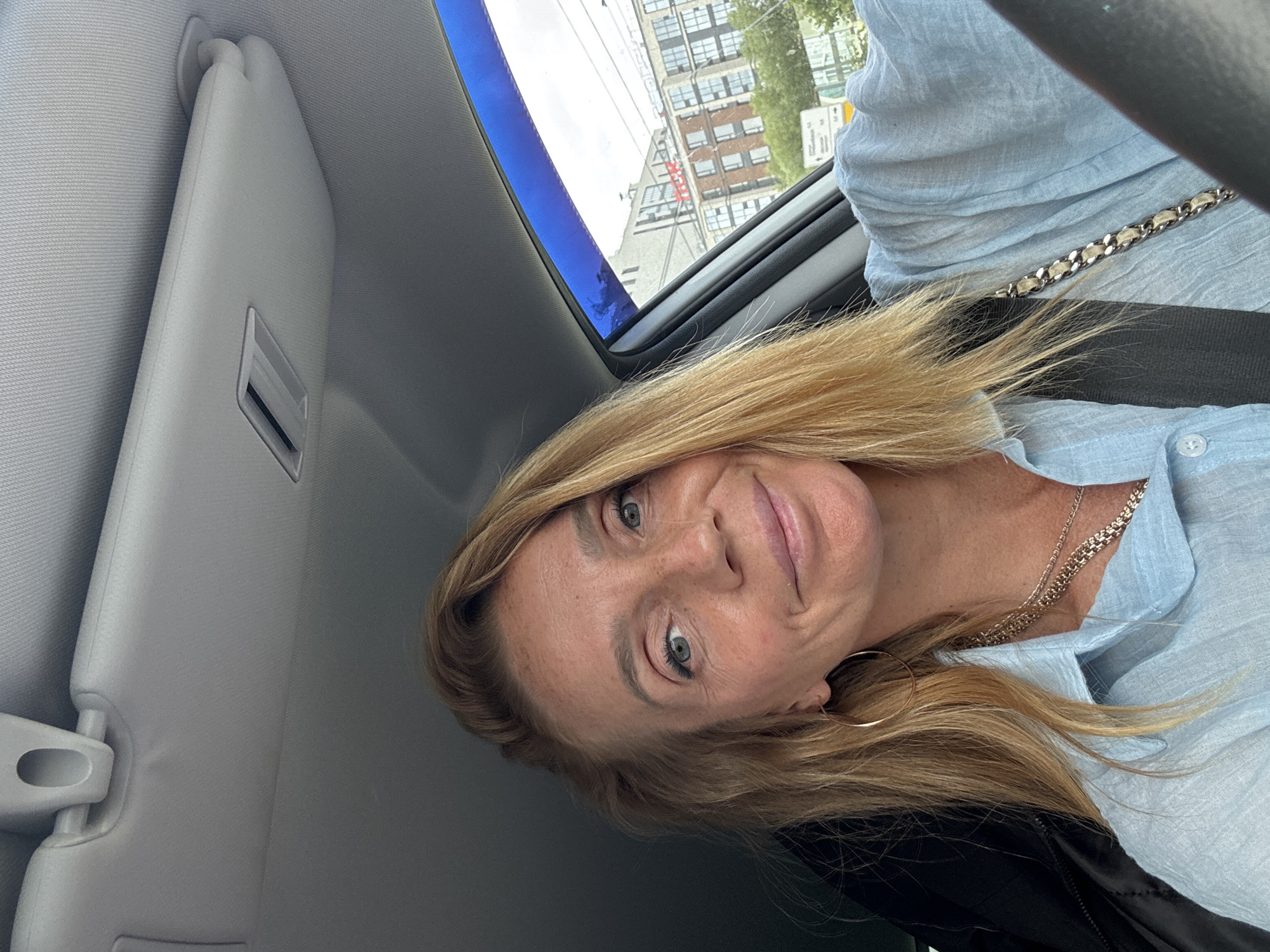 A medium shot captures a fair-skinned woman with blonde hair and blue eyes from the chest up, seated in the driver's seat of a car. She is wearing a light blue button-down shirt, a gold necklace, and a gold hoop earring. Her left hand is on the steering wheel, while her right arm is visible resting on her lap. She is smiling at the camera. The interior of the car is visible, including the sun visor and the passenger-side window. Through the window, a city street with buildings and trees can be seen. The lighting suggests it is daytime.