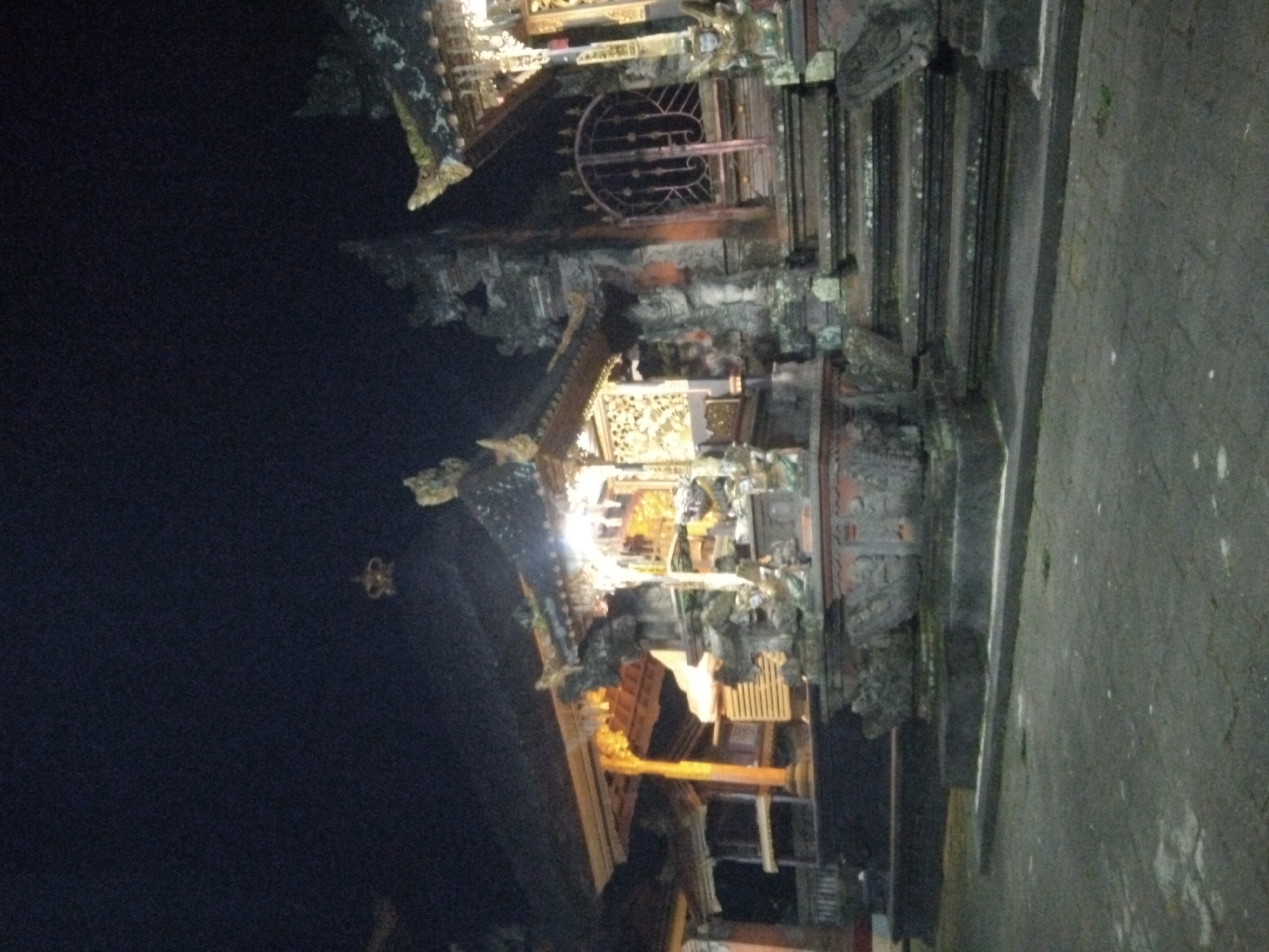 Here's a description of the image:
The image appears to depict a Balinese temple at night. The architecture is ornate, with tiered roofs, intricate carvings, and traditional decorative elements. The temple is lit from within, casting light on its golden details and creating strong contrasts with the surrounding darkness. The foreground shows a paved area, likely a pathway or courtyard adjacent to the temple structure. The overall scene suggests a quiet, atmospheric setting.