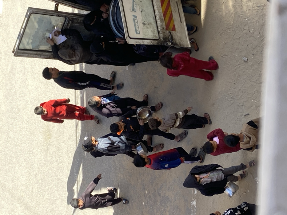 The image depicts an outdoor scene in Khan Yunis, Palestinian Territory, under bright daylight, with distinct shadows cast on a light-colored, possibly concrete or hard-packed dirt, ground surface. Approximately 15-20 individuals, a mix of children, adolescents, and adults, are congregated in the frame. Many are holding empty metal bowls or small pots. On the right side, a white panel van or truck is partially visible with its rear passenger-side door open. The truck's side bears the partially visible text "COVIKAT" and a lower section with diagonal red and yellow stripes. Inside the open truck door, large blue cylindrical containers are visible. One adult near the truck is observed writing on a piece of paper, while another person leans into the vehicle. Several individuals are clustered around the truck, appearing to form a loose line or waiting. A young child in a red jumpsuit stands near the truck, facing it. Other individuals are distributed across the scene, some walking, others standing still, all within proximity to the truck. Clothing varies, including tracksuits, hooded garments, and sandals.