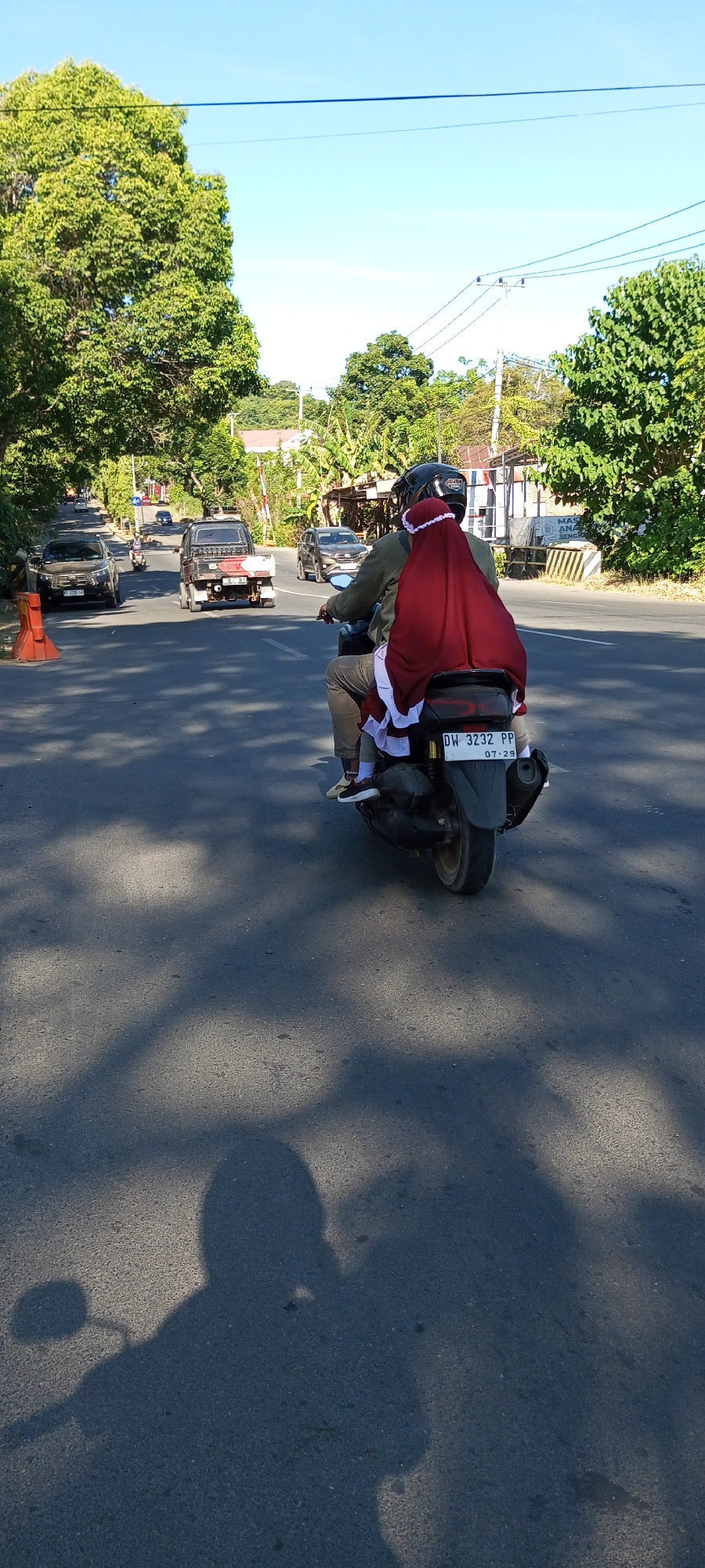 A motorcyclist and a passenger are riding on a paved road in Bulupabbulu, Indonesia, during the day. The motorcyclist is wearing a helmet and a green jacket. The passenger is wearing a maroon hijab and a white and maroon scarf. The motorcycle has a license plate that reads "DW 3232 PP 07-29". In the background, there are trees, other vehicles on the road, and buildings. The sun is casting shadows on the road. The sky is clear and blue.