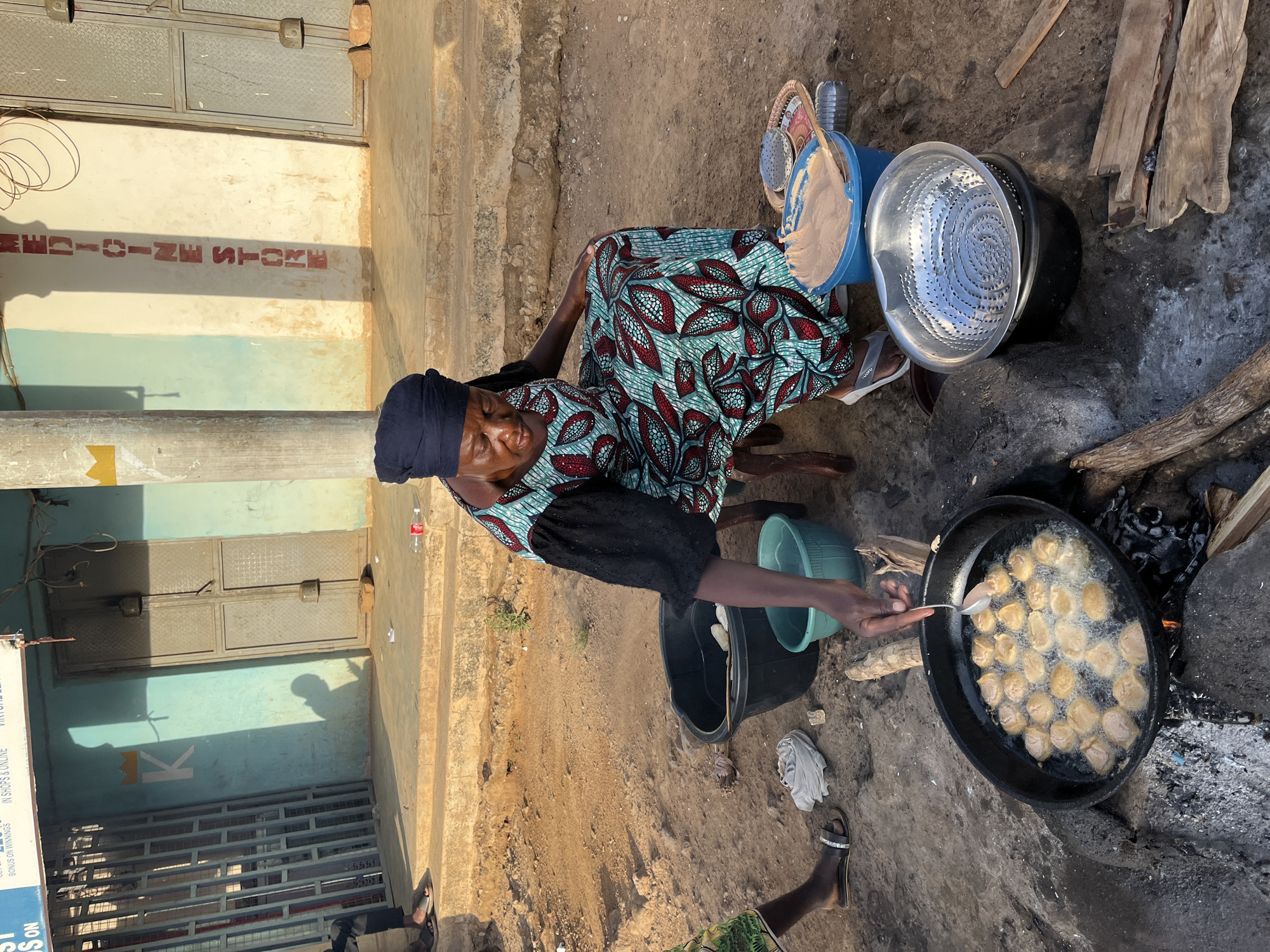 An elderly woman wearing a patterned dress and a head covering is cooking outdoors. She sits next to a large frying pan filled with round, golden-brown fritters, presumably puff-puffs. She is using a spoon to manipulate the fritters in the hot oil. A wood fire burns beneath the pan. Nearby are buckets, a metal colander, and other cooking implements. She appears to be working on a rough concrete surface, perhaps a sidewalk or courtyard. The background includes a building with a blue facade and red writing which says "MEDICINE STORE". Based on information from search results and visual cues in the image, the location is presumed to be in the city of Jos, Nigeria.