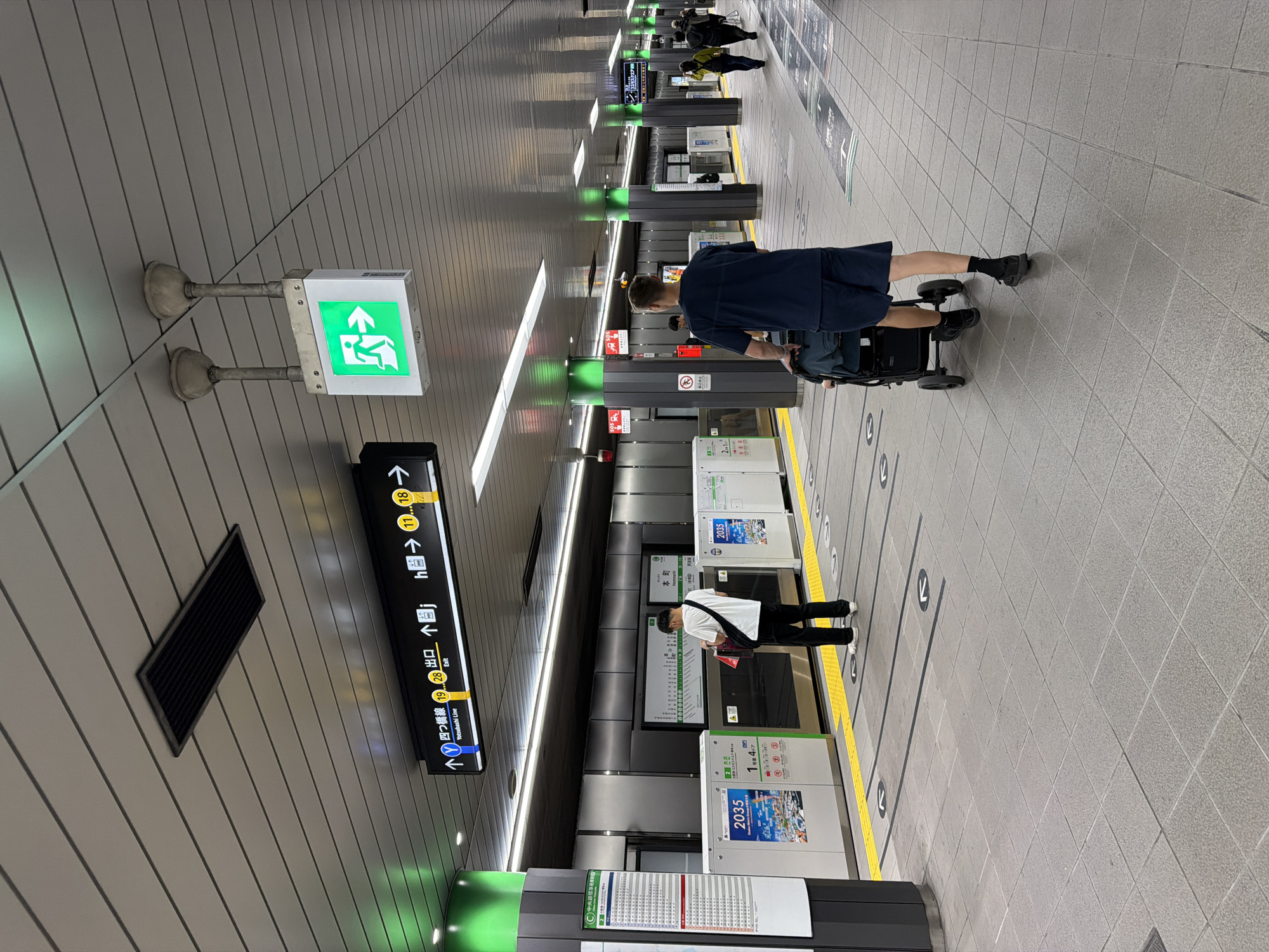 The image depicts a modern, brightly lit subway station platform. The scene is dominated by gray walls, a ribbed ceiling with integrated lighting, and a tiled floor with yellow tactile paving along the platform edge. Black arrow markings on the floor indicate waiting areas.
In the foreground, a person wearing a dark blue shirt and shorts, black socks, and shoes is seen from behind, pushing a black stroller or luggage cart towards the right side of the platform. Further down the platform and to the left, another person in a white t-shirt, black pants, and white shoes, with a black shoulder bag, stands facing an information board. In the background, several other individuals are sparsely distributed along the platform, some appearing to wait.
Notable details include a prominent green illuminated emergency exit sign on the left, featuring a figure with an upward arrow. Overhead, a black directional sign with white and yellow text points to various exits and lines. Visible text on this sign includes "↑四つ橋線出口" (Yotsubashi Line Exit), "Yotsubashi Line," "C中央線" (Chuo Line), "Exit," and exit numbers "19" and "28," along with "本町" (Honmachi). Various station information boards and posters are affixed to pillars and wall panels along the platform, some displaying the number "2035." One poster also shows "1号47." The overall atmosphere is clean and well-maintained, illuminated by bright artificial lights.