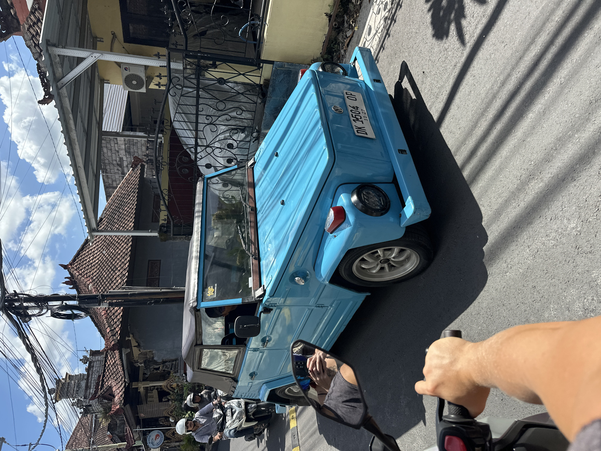 On a sunny day, a bright blue Volkswagen Thing is driving down a paved street in North Kuta, Indonesia. The car, with its top down, has a license plate that reads "DK 1504 OP". A person is riding a motorcycle behind the car, and a glimpse of the rider's arm and shoulder is visible in the foreground. Another motorcycle with two occupants is in front of the blue car. Traditional Balinese buildings with red tile roofs line the street, along with a black metal fence and yellow building. The sky is blue with scattered white clouds, and electrical wires crisscross overhead. Shadows of trees and overhead wires fall on the street.