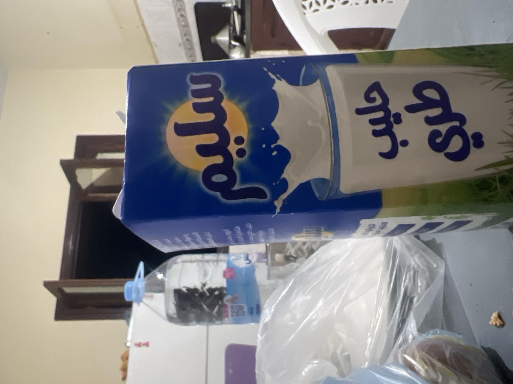 The image shows a carton of "Salim Milk" on a table. The carton is blue and white with Arabic writing. Behind the carton is a plastic water bottle containing some dark objects. There is also a clear plastic bag. The table is gray and there is a piece of food on it. A window is visible in the background. This image cannot be located within the city of Ouaouizert, Morocco.