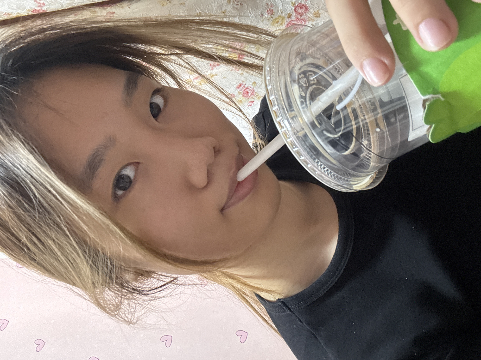 A young East Asian individual with blonde-highlighted brown hair is captured in a close-up, selfie-style shot. They are wearing a black t-shirt and are holding a clear plastic cup with a white straw, from which they are drinking. The background is a pink wall with faint heart illustrations and a floral patterned curtain. The lighting suggests an indoor setting, possibly a home environment in the Shapingba District, China.