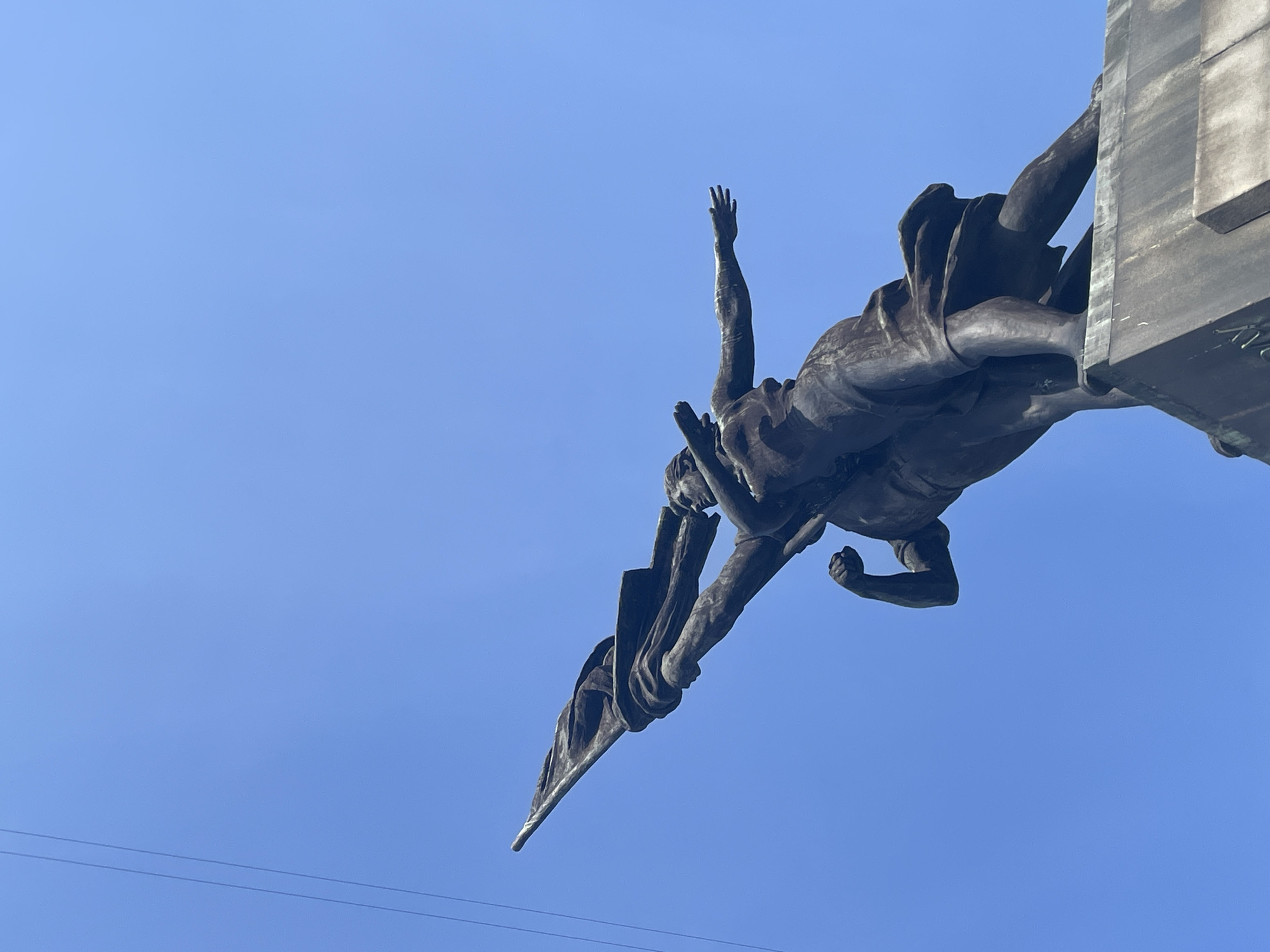A low-angle, upward-facing shot depicts a bronze statue mounted on a concrete plinth against a clear blue sky. The statue features a man with one arm raised in the air and the other holding a flag. The statue is the monument to the heroes of the October Revolution in Yekaterinburg, Russia. Two thin, black horizontal wires can be seen in the bottom left corner of the frame. The statue appears to be well-maintained and securely mounted.