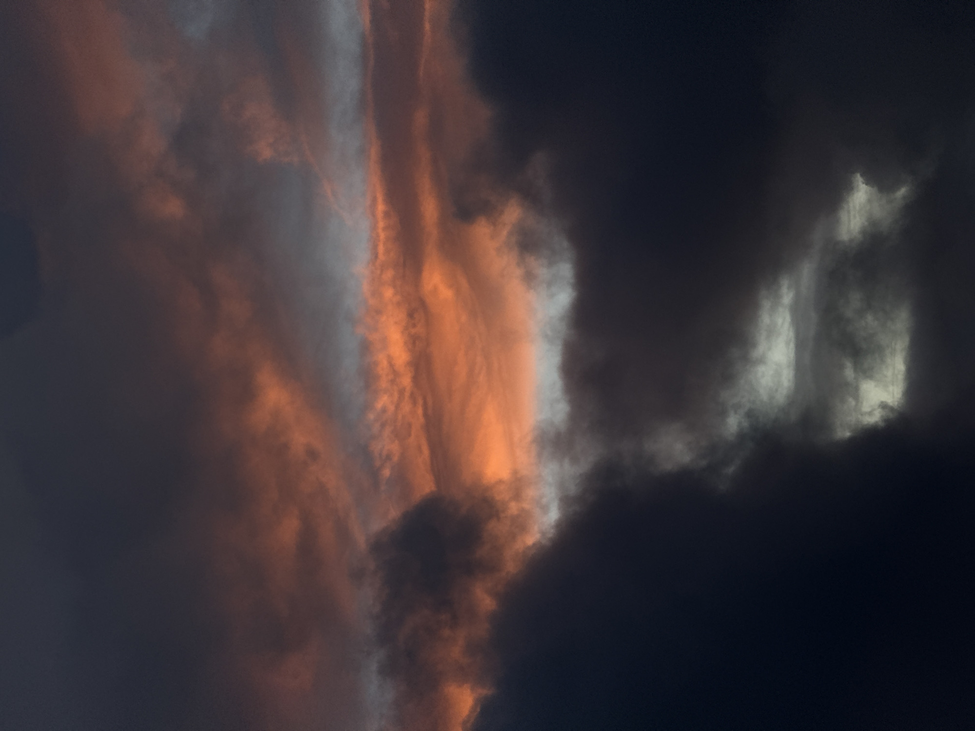 This image captures a dramatic cloudscape over Fécamp, France, likely at sunset or sunrise due to the distinct lighting. The sky is dominated by a striking contrast between brightly illuminated clouds and heavy, dark formations. A prominent vertical band of clouds on the left-center is bathed in vivid orange and reddish hues, with some sections appearing silvery-white. This brightly lit segment stands out against expansive areas of deep blue-grey to almost black clouds, which occupy the right side and other parts of the frame, suggesting a dense cloud cover. The interplay of intense natural light and deep shadow creates a dynamic and powerful visual. There are no people, identifiable objects, or visible text in the image.