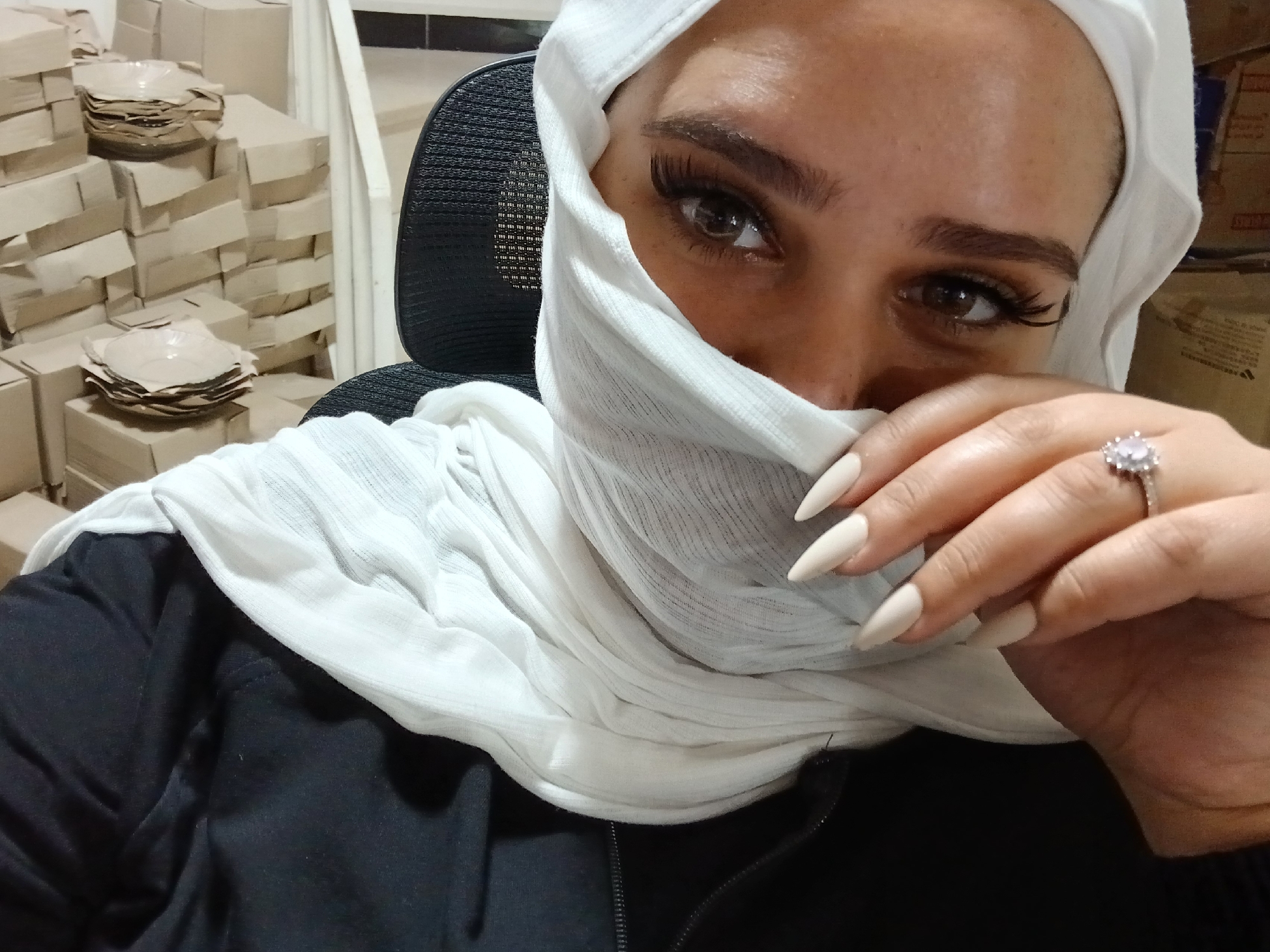 In Rukn ad-Din Municipality, Syria, a close-up, eye-level, medium shot captures a person wearing a white hijab that partially obscures their face. The individual's left eye, adorned with long eyelashes, is visible. Their right hand, with light-colored, pointed fingernails, rests near their chin, and a ring with a light-colored stone is visible on one finger. The person is wearing a black garment. The background is filled with stacked cardboard boxes, some containing stacks of light-colored, circular objects that appear to be bowls or plates. A portion of a black mesh chair is also visible. The scene suggests an indoor environment, possibly a storage area or a workspace.