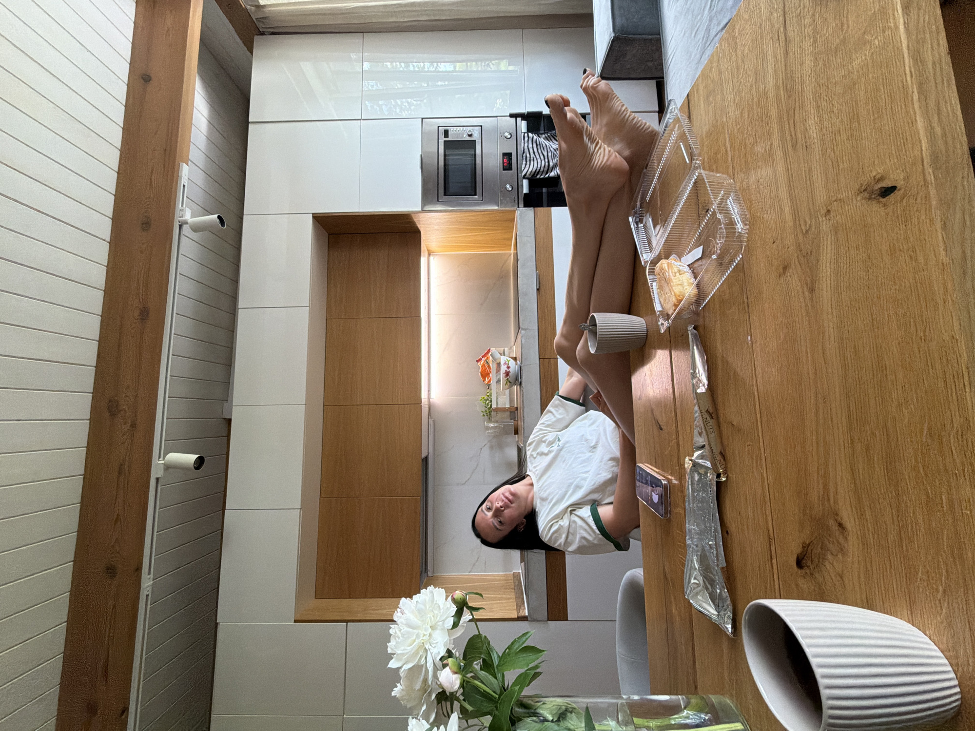 In a well-lit, modern kitchen, a woman is seated at a wooden table with her legs extended. She wears a white t-shirt with green trim. On the table in front of her are a glass vase with white peonies, a phone, a foil-wrapped item, a transparent plastic container with a pastry, a grey ribbed mug, and another mug with a handle. The kitchen features white and wood-grain cabinetry, a microwave, and what appears to be a built-in oven. The ceiling has exposed wooden beams and white paneled sections. The scene is domestic and appears to be a casual moment. The city of Chaikovsky, Russia is not depicted in this image.