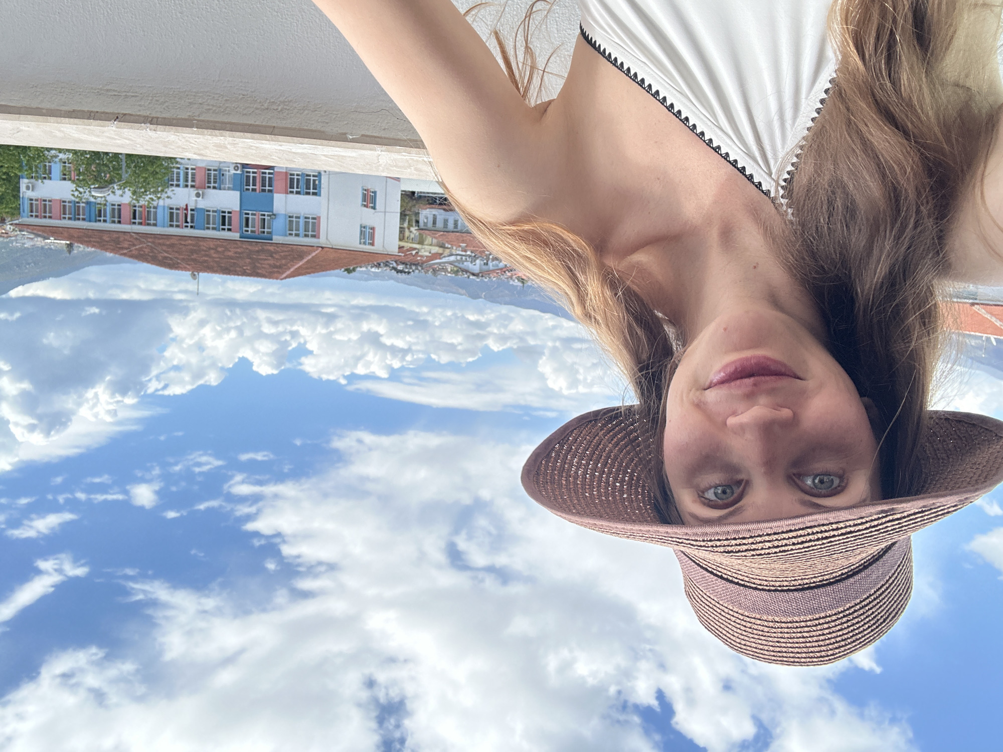 A woman wearing a straw hat and a white top is visible against a backdrop of a building, mountains, and a cloudy sky. The building appears to be a school, identifiable by its blue and white exterior with pink accents. The mountains are in the distance, and the sky is filled with white clouds. The image appears to have been taken upside down, with the woman's face in the foreground. The location is Fethiye, Turkey.
