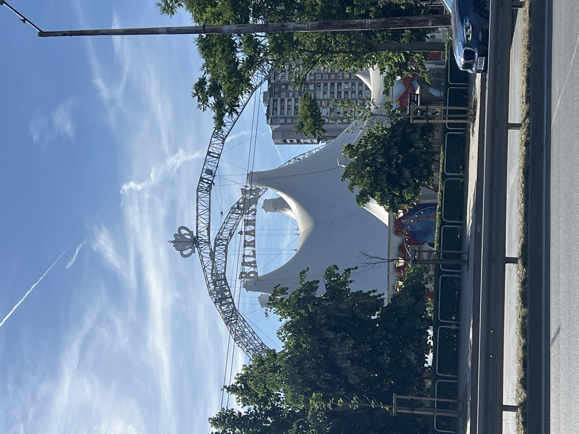 The image shows the "Balkanski" circus tent in Sofia, Bulgaria. The white tent structure is visible, as well as the metal frame with the circus' name. A building can be seen behind the tent. There are trees and greenery in the foreground. A road with a vehicle parked on the side is visible on the right. The sky is blue with some clouds.