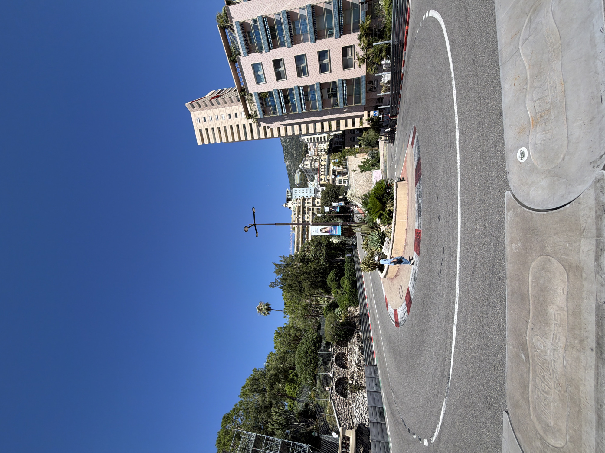 The image shows a sunny day in Monaco, likely during a daytime event or before one begins. The scene depicts a famous section of the Monaco Grand Prix circuit, specifically the hairpin turn near the Fairmont Hotel, though the specific turn is not explicitly identifiable. The race track is asphalt, marked with white lines. Red and white curbing lines the sides of the track. In the foreground, a man in a light blue shirt and dark pants is walking away from the camera, having just passed a small circular garden island in the middle of the turn. This island features greenery and some cacti.
To the left of the track, there's a sloping hillside covered in lush green trees and vegetation. A stone wall with arched structures and metal fencing is visible along the hillside. Scaffolding is erected on the far left, suggesting ongoing construction or preparation. On the right side of the track, a pink apartment building with balconies and blue awnings dominates the view. Behind it, another taller, pale building is visible, with a curved top. The sky is a clear, bright blue. In the very foreground, a concrete structure with an imprint resembling a shoe or footprint is visible, with some text that is partially legible, possibly referring to "RECARO" or "RECAPRO". There are also some banners visible along the trackside.