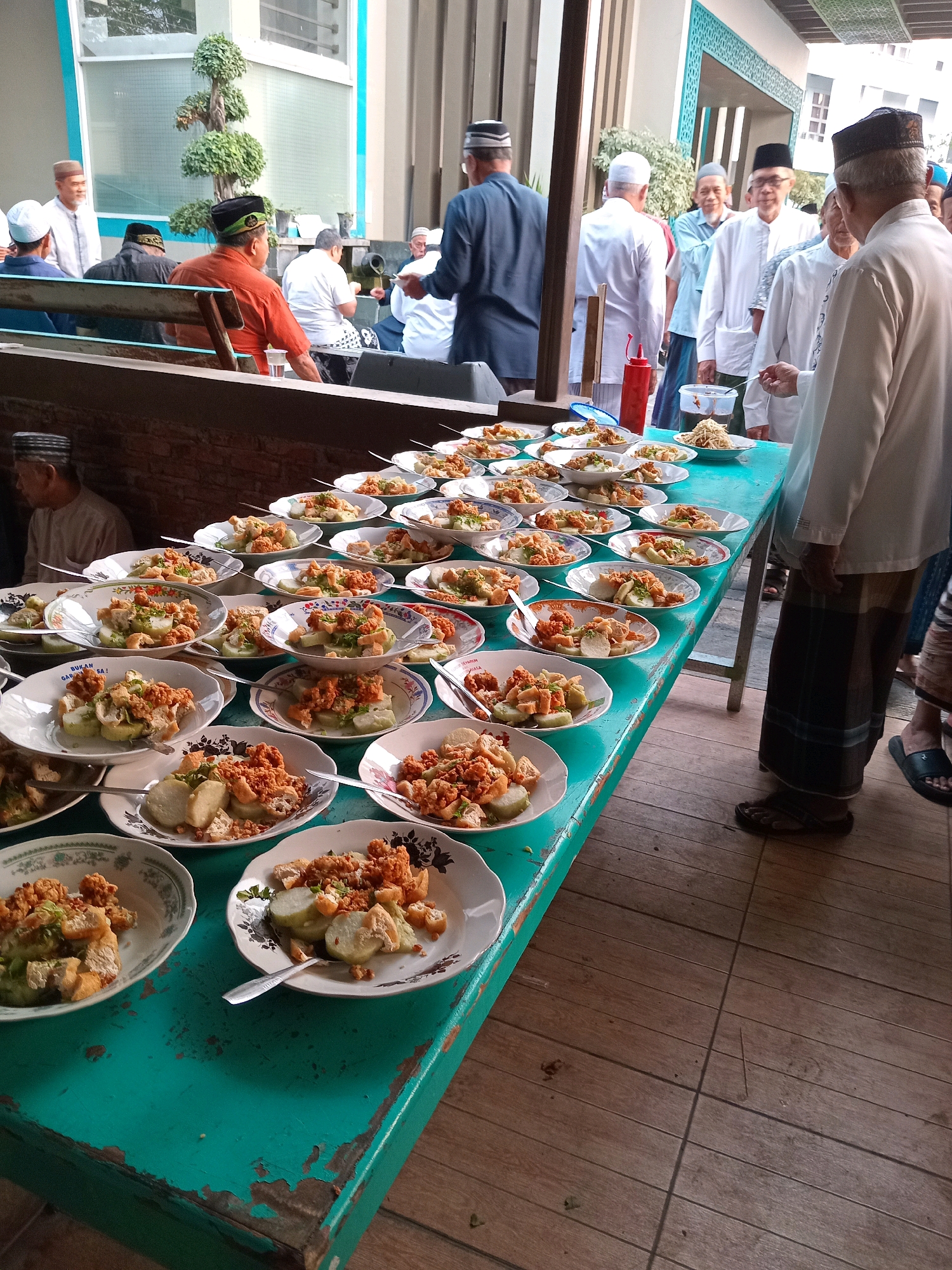 This image depicts an outdoor gathering at a location in Randuagung, Indonesia, likely a mosque or community center, given the presence of men in Islamic attire and the architectural style. The scene is centered around a long, turquoise-colored table laden with numerous plates of food. These plates, mostly white with floral or patterned rims, are arranged in neat rows and contain a dish composed of white, rectangular and spherical food items topped with a crumbled, reddish-brown substance and green garnishes.
Several individuals, predominantly men, are visible in the frame, many wearing peci caps and traditional Indonesian clothing. Some are standing, observing the food, while others are seated on benches or on the ground in the background, engaging in conversation or waiting. The food appears to be prepared for a communal meal or event, possibly a religious celebration or a community gathering. The backdrop includes a brick wall, a building with large windows, and some greenery, suggesting an established outdoor space. The lighting indicates it is daytime, with natural light illuminating the scene. The overall atmosphere suggests a social or religious event where food is being served to a group of people.