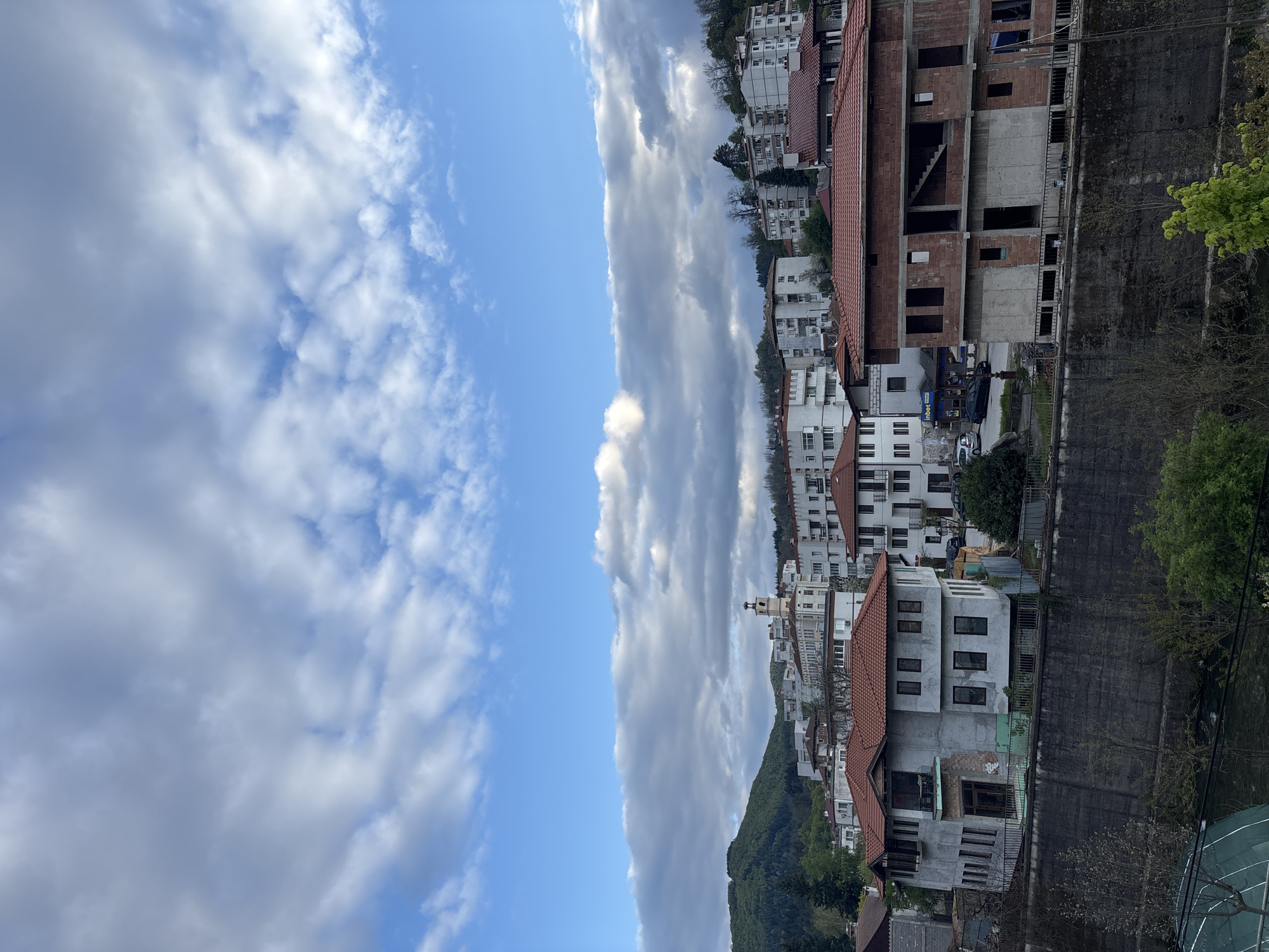 The image depicts a view of Zlatograd, Bulgaria, showcasing a mix of residential buildings with red-tiled roofs and a taller, unfinished brick and concrete structure. The architecture is clustered along the right side of the frame, adjacent to a retaining wall. A green hillside with trees and some additional houses is visible in the lower part of the frame. The left half of the image is dominated by a sky filled with white and gray clouds against a blue backdrop. There are no people visible. A blue car is seen next to the unfinished brick building.