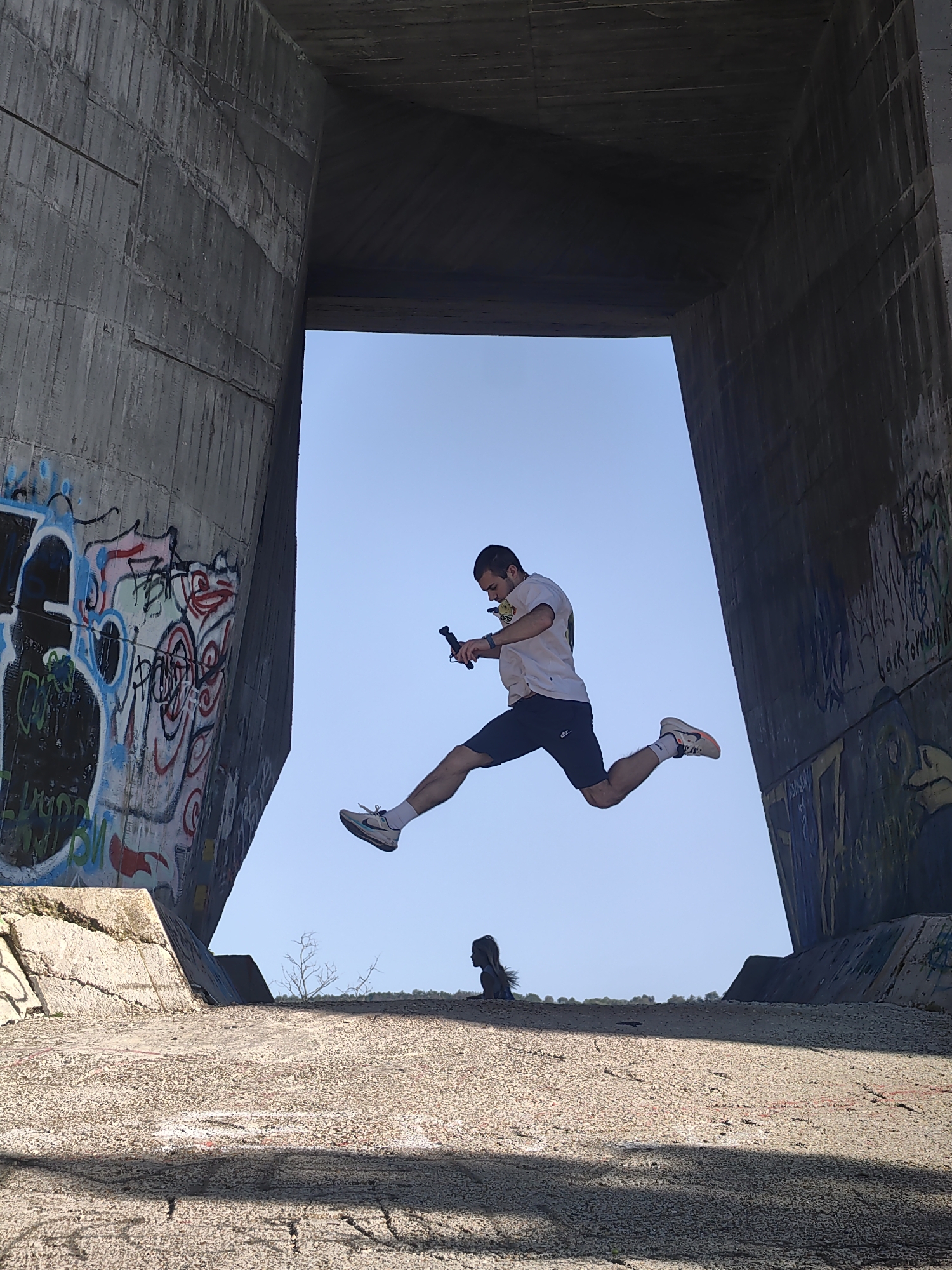 A man is photographed mid-air, jumping between two concrete structures. He wears a white t-shirt, dark shorts, and white shoes with a dark swoosh. He appears to be holding a dark object in his right hand. In the background, a girl is visible standing on the ground between the concrete structures, silhouetted against the sky. The concrete structures are covered in graffiti. The scene is lit by bright daylight. The location is in Varna, Bulgaria.