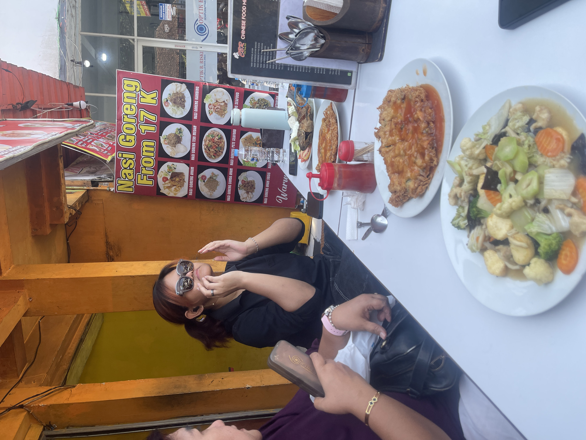 Two women are seated at a white table outdoors in front of a building with an orange facade. The woman on the left is partially visible, wearing a purple shirt and gold bracelets. The woman on the right is wearing a black collared shirt and sunglasses, with her hands near her face. On the table are plates of food, including fried rice and a vegetable stir-fry, as well as condiments and cutlery. A sign behind the women advertises "Nasi Goreng From 17 K". The environment suggests a restaurant in Pacet, Indonesia, based on the signage.