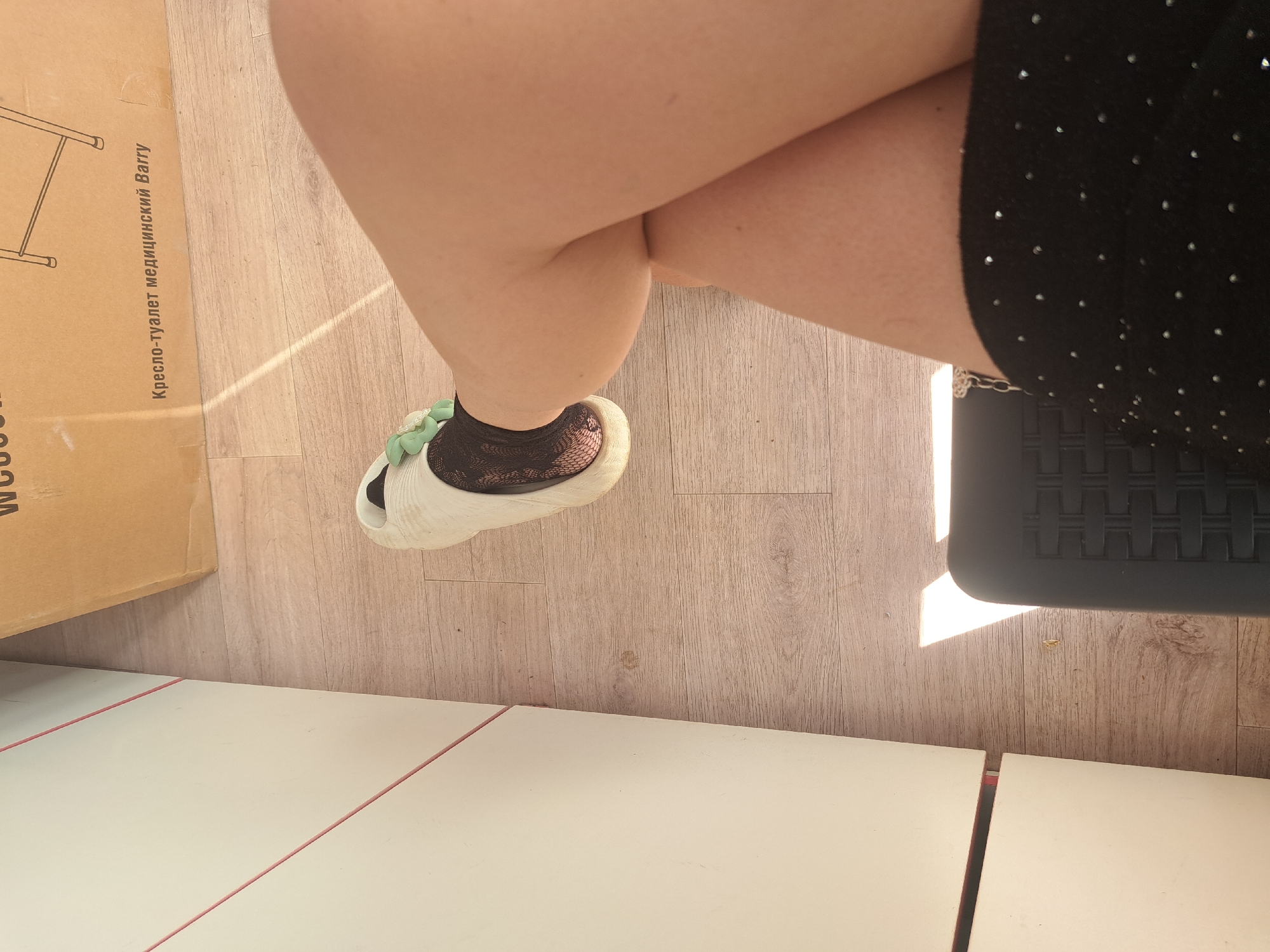 Here is a forensic description of the image:
The image is a high-angle shot showing a partial view of a person sitting with crossed legs. The person is wearing a short, dark skirt with light-colored dots and a pair of white sandals with a decorative green bow. A patterned, possibly crocheted, sock is visible inside the left sandal. The person is sitting on a black chair with a slatted seat. To the left is a cardboard box with Russian text, including the words "Кресло-туалет медицинский Ваггу". The surface beneath the box and chair appears to be wooden flooring. In the foreground is a white surface with a red line along its edge, possibly a table. The location is likely in Leningradskaya, Russia based on the Russian text on the box. The image depicts a casual scene indoors, possibly a home or medical setting given the text on the box.