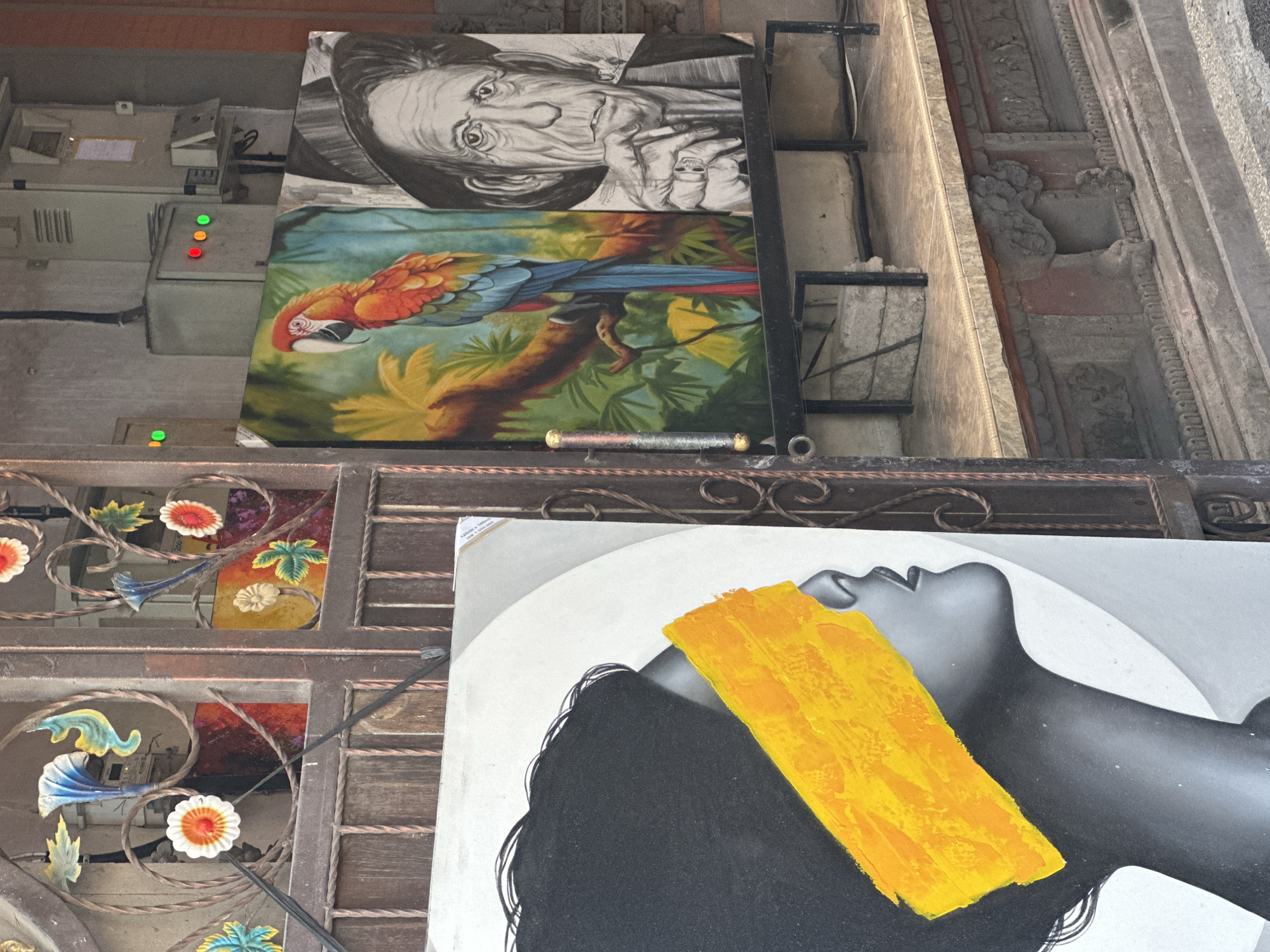 An outdoor art display is set up on a street in Ubud District, Indonesia, in front of a building with electrical boxes and some decorative metalwork. The display features three canvases. On the left, a large black and white painting depicts the profile of a woman with dark hair and a bright yellow band across her eyes. To the right, two more canvases are presented side-by-side. The left canvas shows a colorful parrot perched on a branch, surrounded by lush green foliage. The right canvas is a black and white sketch of an older man wearing a hat, with his hand near his mouth. The paintings are displayed on a black metal stand, with the larger canvas leaning against a metal gate structure. The background shows a textured wall and some decorative architectural elements.