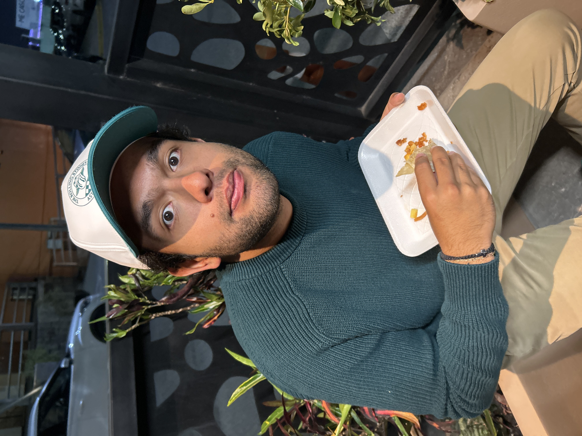 A man with a dark beard and wearing a white and green baseball cap and a dark green sweater is holding a white foam container of food. He is looking at the camera with a slightly silly expression, his tongue sticking out slightly. He is wearing light tan pants and sitting outside. Behind him is a black fence with green plants. A car is visible behind the fence. The location is Unknown, Unknown.