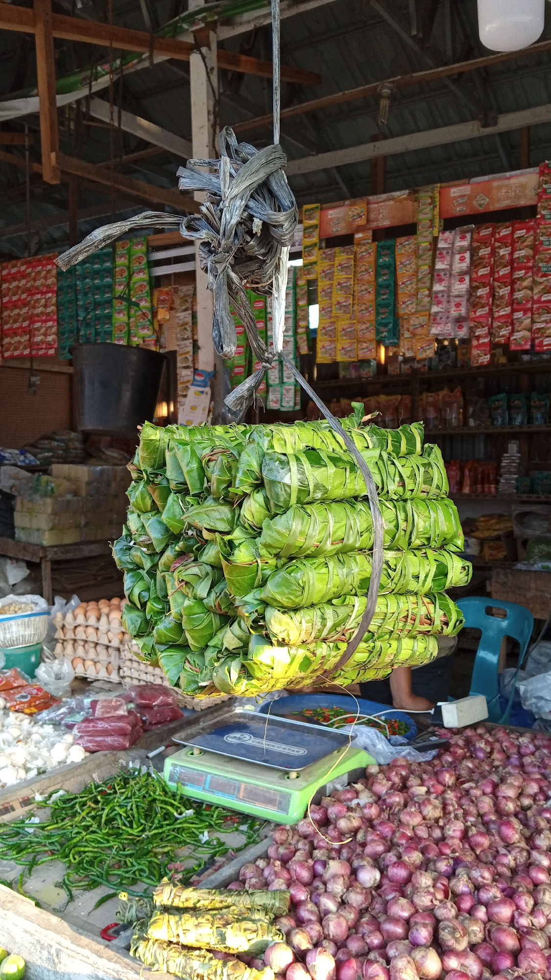 This image depicts a bustling outdoor market in Puloara, Indonesia. The central focus is a large, horizontally-oriented stack of food items, possibly rice or snacks, wrapped in green leaves and tied with rough string, suspended from the ceiling by a thick rope. The market is set up under a rustic wooden and corrugated metal roof structure, with shelves in the background displaying an assortment of packaged goods, primarily in brightly colored wrappers.
In the foreground, the market stalls are laden with fresh produce. Piles of red onions and long green chilies are prominently displayed. To the left, there are groupings of garlic and several packages of raw red meat. A green and white analog scale sits on a wooden surface, indicating ongoing transactions or preparation for them. A blue plastic chair is visible in the mid-right background, and a person's arm and shoulder can be seen seated behind it. The overall impression is one of a vibrant and active marketplace, typical of local Indonesian commerce.