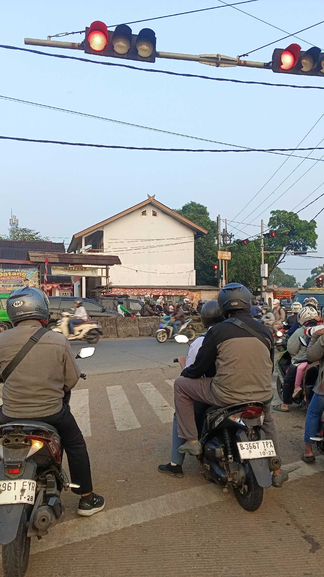 The image captures a traffic intersection in Kemang, Indonesia, likely during daylight hours given the illumination. Multiple traffic lights are visible, with at least one displaying a red signal, indicating a stop. Several motorcycles, some with riders and some with passengers, are stationary at the intersection, suggesting they are obeying the traffic signal. The foreground shows the backs of two motorcycles and their riders, who are wearing helmets for safety. The motorcycles are parked on a paved road with visible pedestrian crossing markings. In the background, a white building with a distinctive pitched roof stands prominently, flanked by trees and other structures. Various banners and signs are present on buildings and poles, contributing to the urban environment. The overall scene depicts a typical bustling intersection in an Indonesian city.