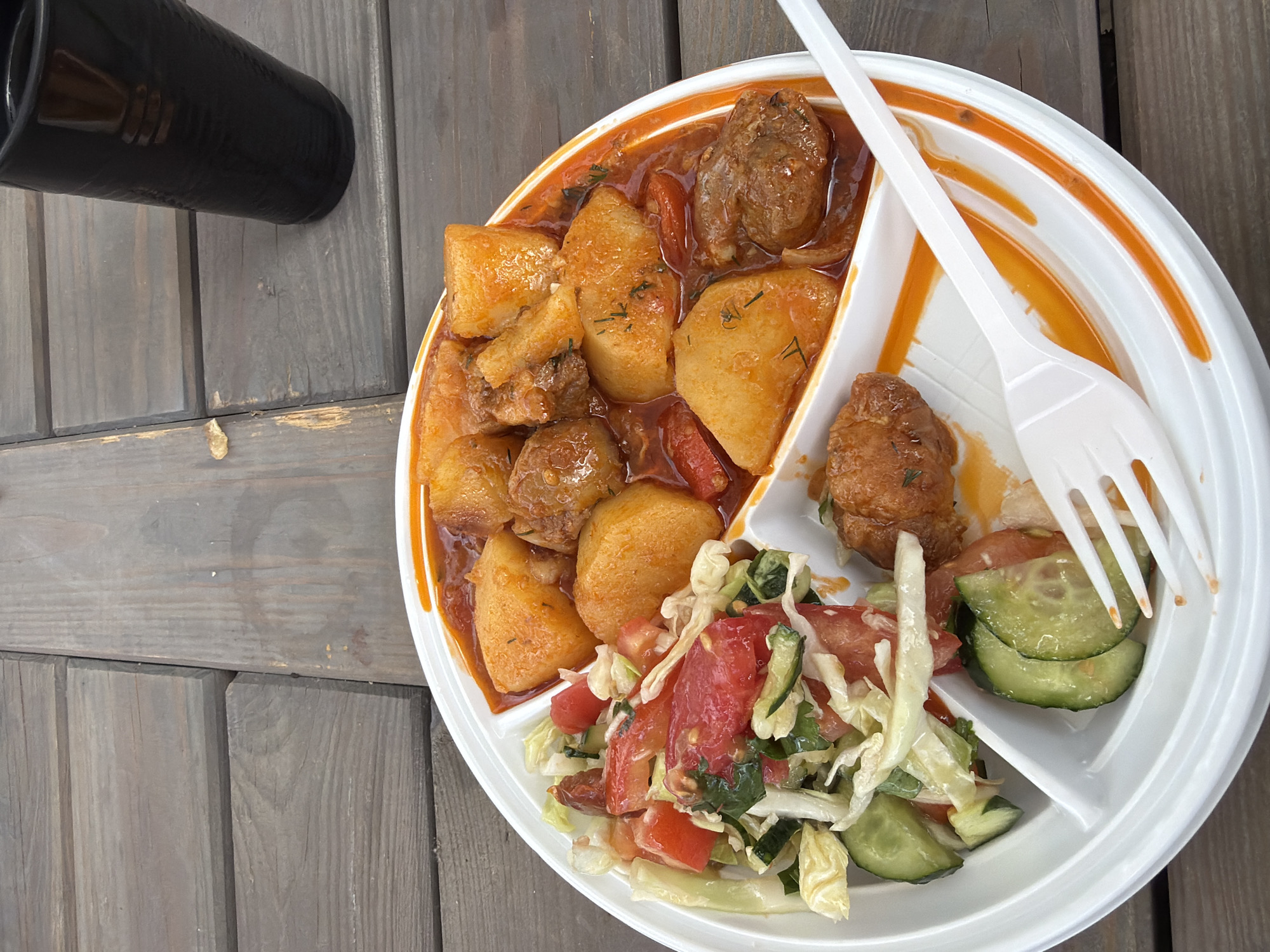 A top-down, close-up shot shows a segmented white plastic plate of food sitting on a weathered wooden surface. To the left of the plate is a black cup. The plate has three sections: One section contains a stew with large pieces of potato and meat in a reddish sauce. Another section holds two meat pieces with the same reddish sauce. The third section contains a salad with tomatoes, cucumbers, and shredded cabbage. A white plastic fork and spoon rest on the plate. The location is Khimki, Russia.