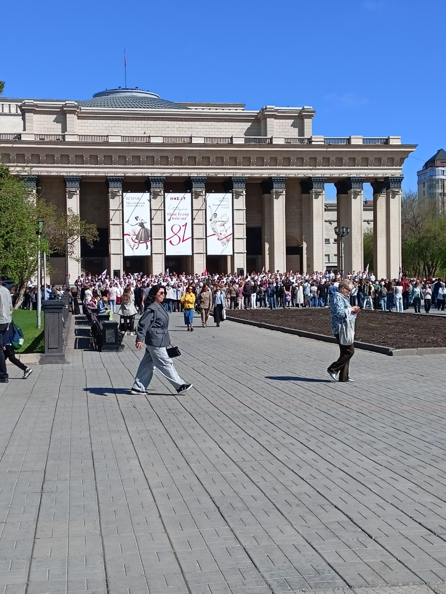 The image depicts a wide-angle shot of a public square in Novosibirsk, Russia, with a large, classical-style building dominating the background. The building has a series of columns supporting a horizontal entablature. Banners are hung between the columns displaying text and ballet-related imagery.
In the foreground, a paved walkway leads towards the building. Two women are walking across the walkway, one wearing a gray suit with matching pants and the other wearing a light blue patterned jacket with brown pants. To the left, there are benches and a small grassy area with trees. A crowd of people stands in front of the building. The sky is clear and blue.