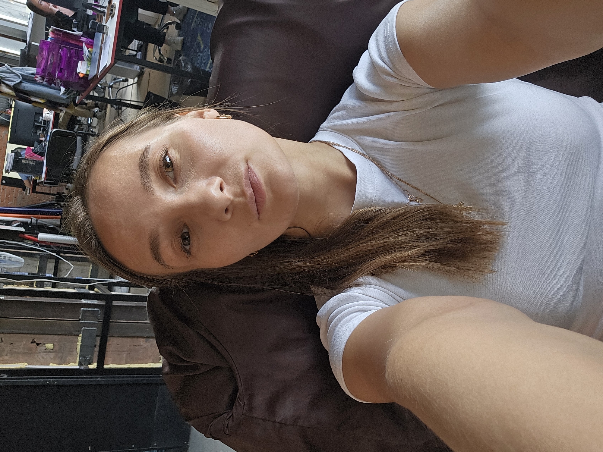 A woman is lying on a dark brown couch, presumably indoors. She is wearing a white t-shirt and a gold necklace. Her brown hair is partially draped over the couch. Her face is visible, and she appears to be looking at the camera. In the background, there is a cluttered desk with various items, including what appears to be office supplies and electronic devices. The location is indiscernible without further visual cues.