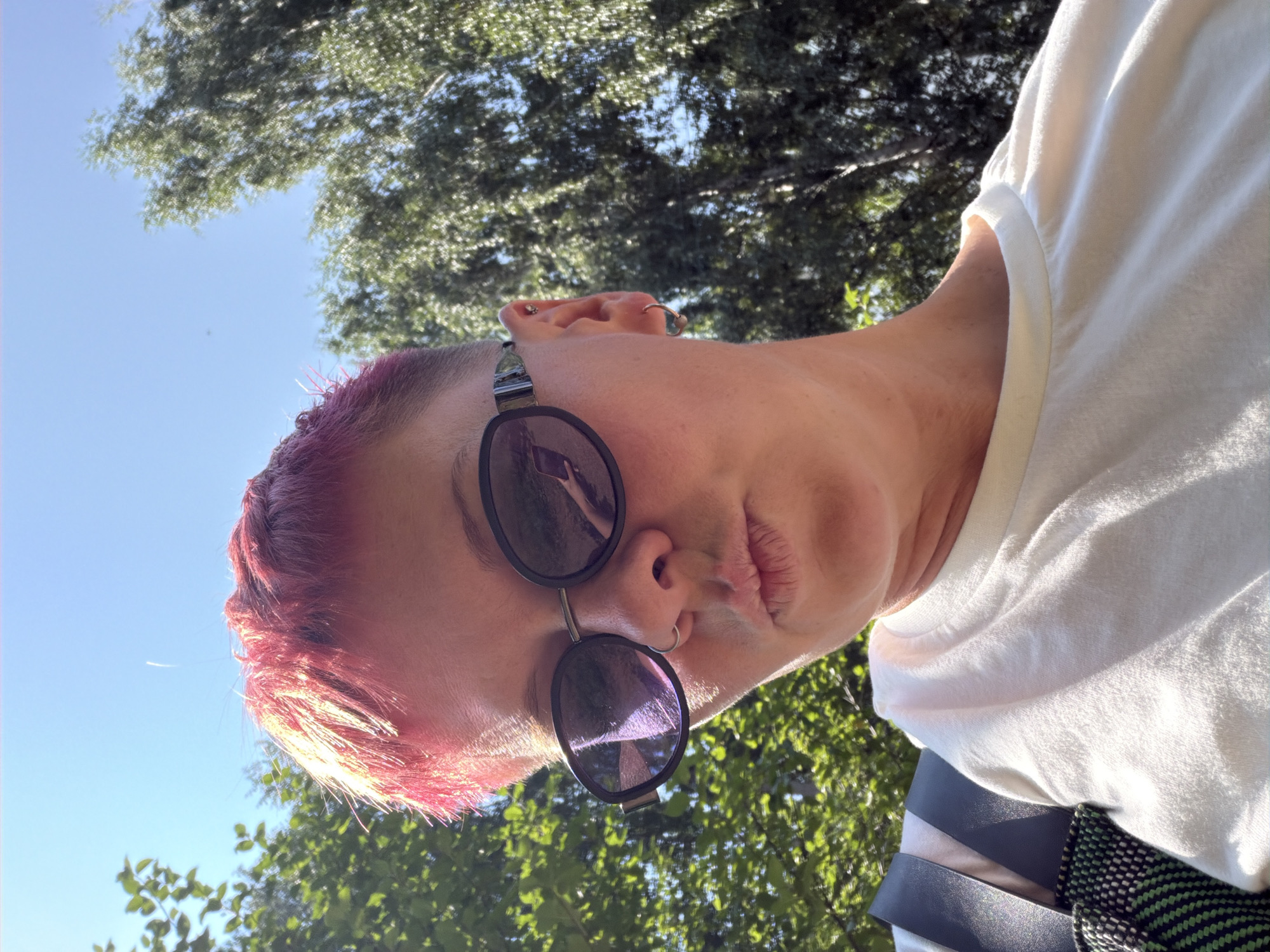 A close-up, low-angle selfie captures a person with short, brightly dyed pink hair, wearing round, dark-lensed sunglasses and a plain white t-shirt. They have a nose ring and an earring on their visible ear, and their lips are pursed. The background consists of lush green trees and a bright blue sky, suggesting an outdoor setting. There is a strap from a bag visible on the left side of the frame. The image appears to have been taken in Chaikovsky, Russia, as indicated by the provided context.