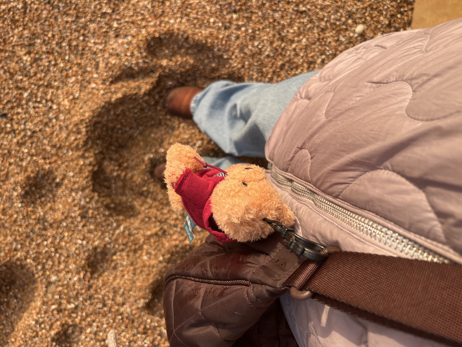 The image displays a top-down perspective of an individual standing on a shingle beach consistent with the coast near Swyre, United Kingdom. The ground surface consists of tightly packed small, irregularly shaped light brown and tan pebbles. The subject is observed wearing light blue denim jeans and brown shoes. A light purple quilted jacket with a visible silver zipper runs vertically up the center. A dark brown quilted bag, equipped with a wide brown fabric strap, is positioned on the left side of the frame. Clipped to the bag with a black carabiner-style clasp is a small, light brown teddy bear plushie, which wears a red top and has a small tag attached to its back. Direct sunlight casts distinct shadows onto the pebbled beach surface.