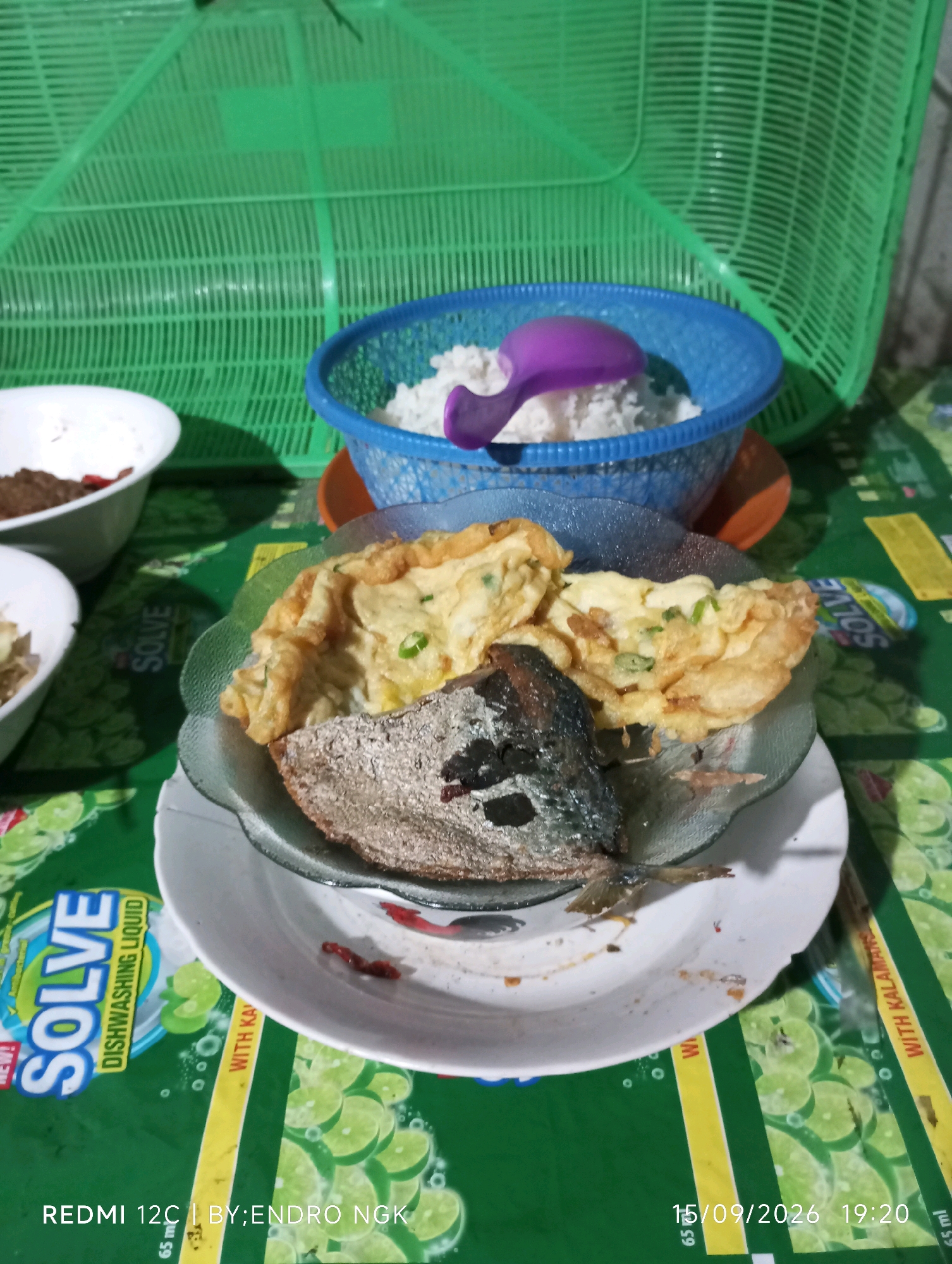 A close-up, eye-level, horizontal shot captures a simple meal laid out on a green tablecloth patterned with sliced limes. In the foreground, a white plate holds a piece of fried fish, appearing slightly charred, alongside a fried egg omelet with visible green onions. To the left, two small white bowls contain what appears to be food. Behind the plate, a blue plastic basket overflows with white rice, and a purple plastic spoon rests on top. In the background, a large, green plastic basket with a mesh pattern looms over the scene. The image appears to have been taken indoors, likely in a residential setting in Ringinanom, Indonesia, given the presence of the "REDMI 12C | BY; ENDRO NGK" watermark, suggesting the photo was taken by a local individual with that device and potentially in that area. The lighting is dim, suggesting it might be evening or a poorly lit interior. The overall impression is one of a humble, home-cooked meal.