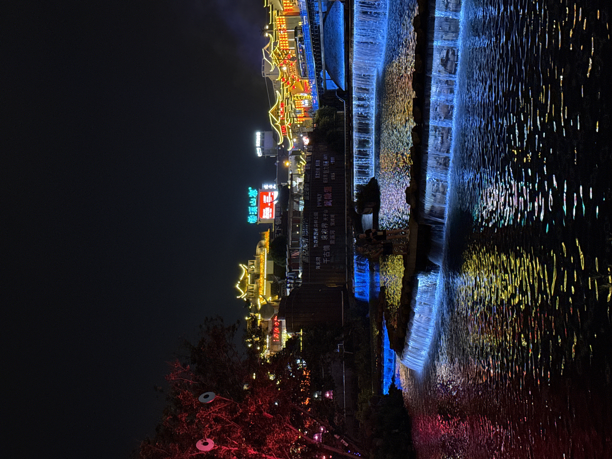 The image depicts a night scene in Wulingyuan, China, featuring a water feature illuminated by blue and yellow lights. To the left, a cityscape with traditional Chinese architecture is visible, with buildings adorned with yellow lights. A tree with red lights stands in the foreground. Signs in Chinese are displayed on the buildings, including "崇溪山寨 (Chongxi Mountain Village)" and "张家界国家森林公园 (Zhangjiajie National Forest Park)". The water feature is cascading in steps, reflecting the colorful lights. The overall ambiance is vibrant and illuminated.
