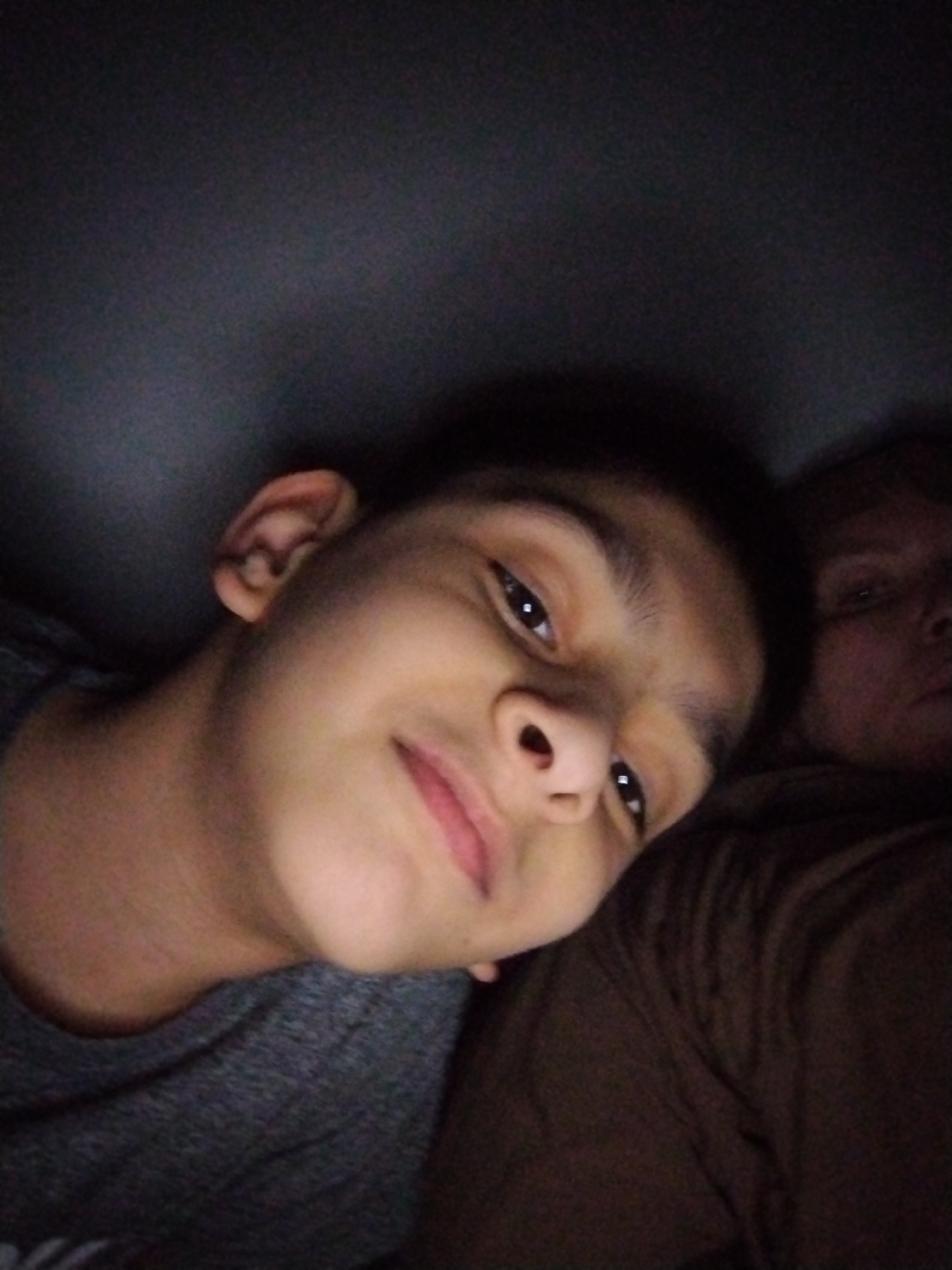The image shows a close-up, slightly distorted selfie of two individuals. The person in the foreground, on the left, is a young male with dark hair and brown eyes. He is wearing a dark grey t-shirt and appears to be smiling, with his mouth slightly open. His head is tilted, and his left ear is visible. To the right and slightly behind him is another person, partially visible, whose features are obscured by low lighting and distance. This second individual appears to have lighter skin and is wearing a brown garment. The background is dark and indistinct, suggesting an indoor or dimly lit environment. There is no discernible indication of the city of Al Mas'oudi, United Arab Emirates, within the image itself.