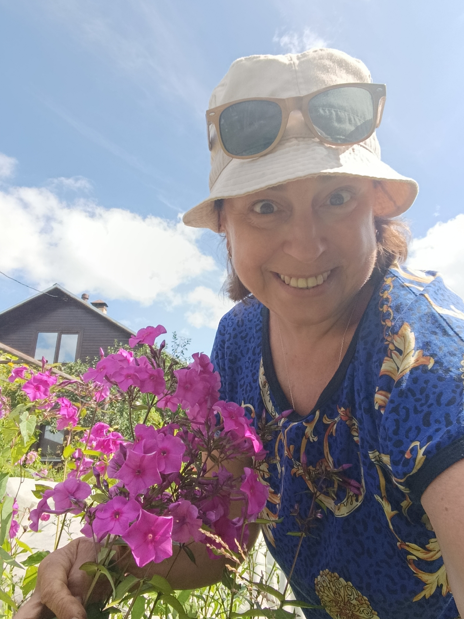 An outdoor, eye-level, medium shot captures a woman holding a bouquet of vibrant pink flowers in the foreground. She is wearing a beige bucket hat and sunglasses, and a blue t-shirt adorned with a gold and black ornate pattern. The woman is smiling and looking directly at the camera, with her eyes wide open. Behind her, a dark wooden house with a brown roof and a chimney is visible, set against a bright blue sky with scattered white clouds. Lush green foliage and more pink flowers surround the woman and the house. The scene appears to be in a garden or yard, possibly in the city of Vereya, Russia, given the architectural style of the house and the type of vegetation.