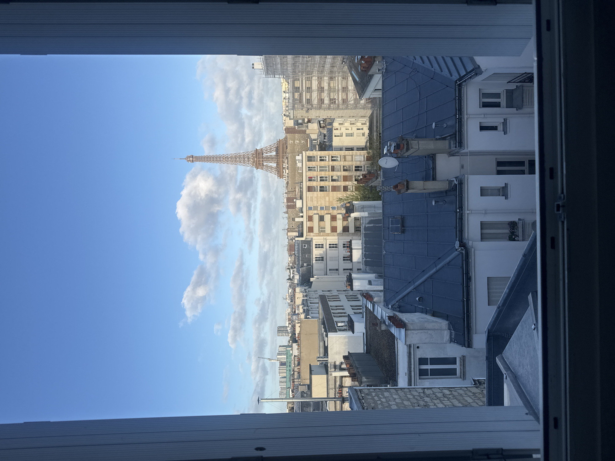 The image shows a view of Paris from a window. The Eiffel Tower is prominently visible in the distance, flanked by buildings. The sky is blue with scattered clouds. Closer to the viewer, the scene includes the rooftops of Parisian buildings, some with blue roofs. The perspective is from inside a building, looking out through a window frame.