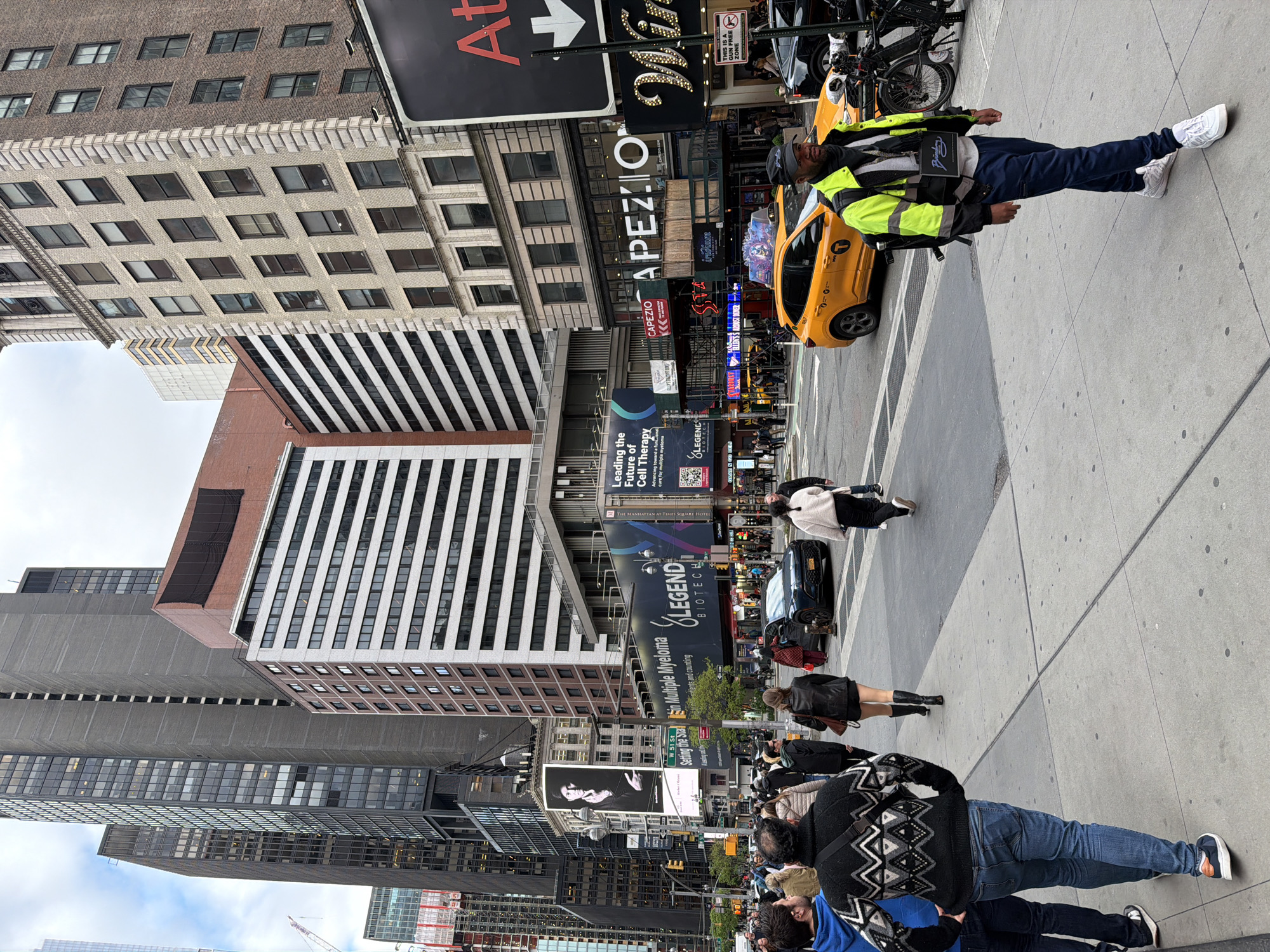 This media file captures a vibrant daytime street scene in New York City, likely near Times Square, as indicated by the "THE MANHATTAN AT TIMES SQUARE HOTEL" sign and "W 51 St" street sign. The perspective is looking down a busy street lined with numerous tall buildings of various architectural styles, including brick facades, concrete structures, and modern glass-windowed skyscrapers. The sky appears overcast.
In the foreground and midground, there are many pedestrians on the sidewalk. A man in a high-visibility neon yellow vest, dark pants, and white sneakers stands prominently on the right side of the sidewalk, appearing to be walking or waiting. A yellow New York City taxi cab is partially visible behind him on the street, and other vehicles can be seen further in the distance. People are scattered across the foreground and midground sidewalks, engaged in daily activities.
The buildings are adorned with large, illuminated advertisements and digital billboards. Visible text includes "CAPEZIO" (a dancewear company), "LEGEND BIOTECH" promoting "Leading the Future of Cell Therapy" and "Advancing toward a functional cure for multiple myeloma," and a sign that reads "THIS IS A GUN FREE ZONE." Other signs show "GRAB & GO DELI" and a large "At" sign with an arrow. The scene depicts a typical bustling urban environment with a mix of commercial activity and pedestrian traffic.