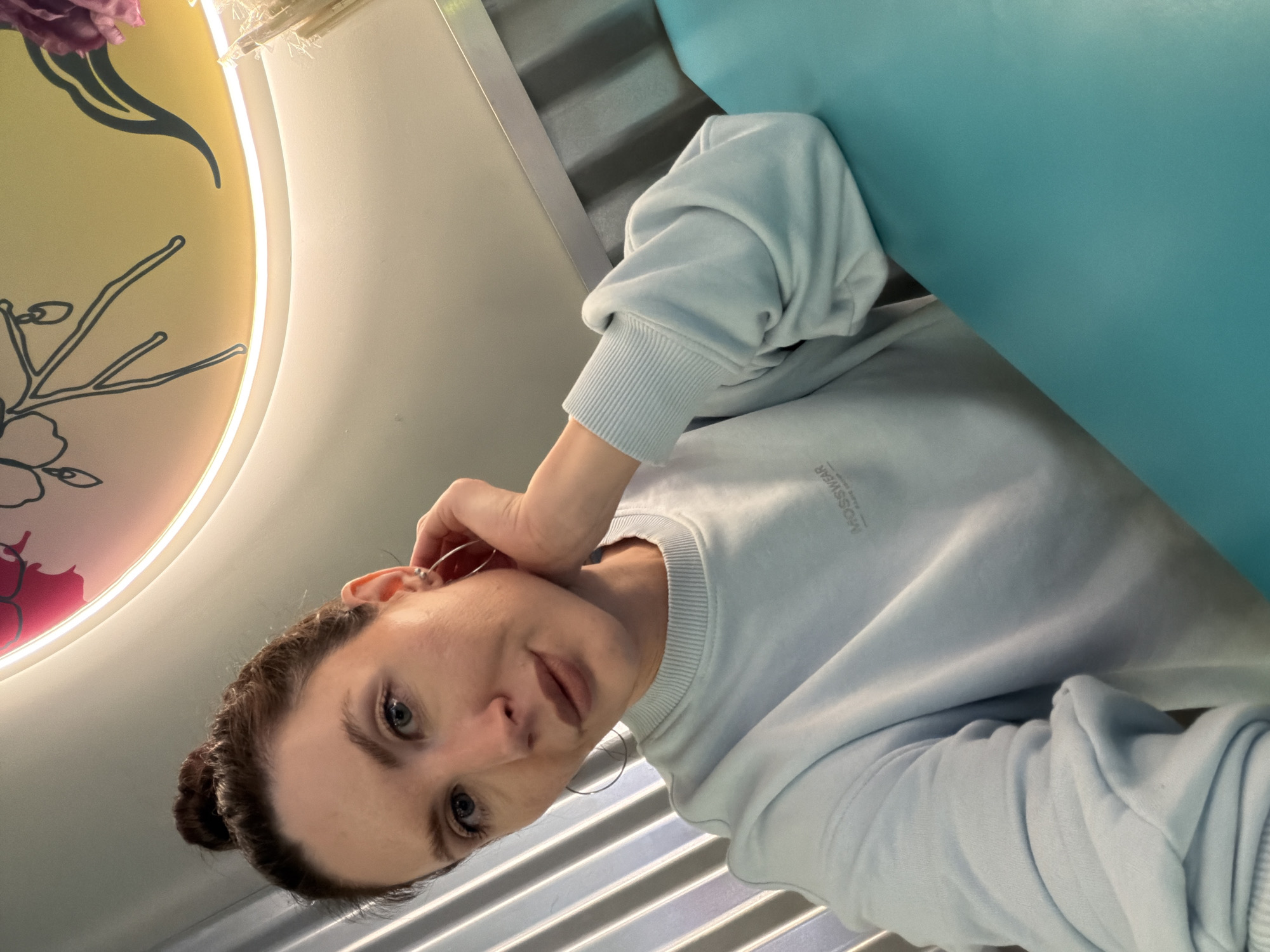 The image depicts a woman in a light blue sweatshirt. She is seated next to a teal-colored wall. A decorative circular light fixture with floral designs is visible in the background. The photo was taken inside the restaurant "Sashas restaurant" in Perm, Russia.
