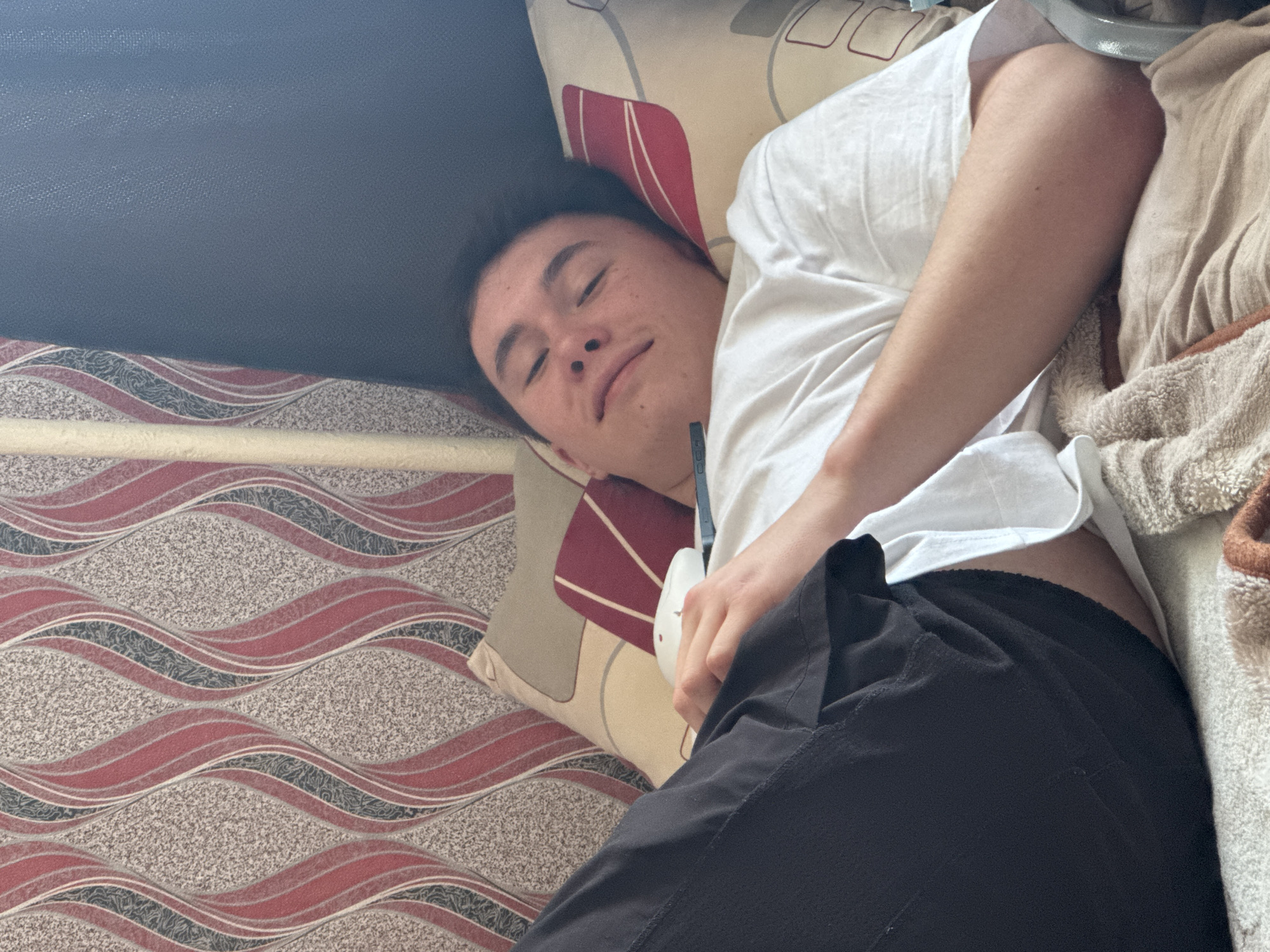 A young man with dark hair lies on a bed with a pillow featuring red and beige designs. He is wearing a white t-shirt and black pants. He is holding a white object in his hand. The bed is positioned against a wall with patterned wallpaper. The location is Perm, Russia.