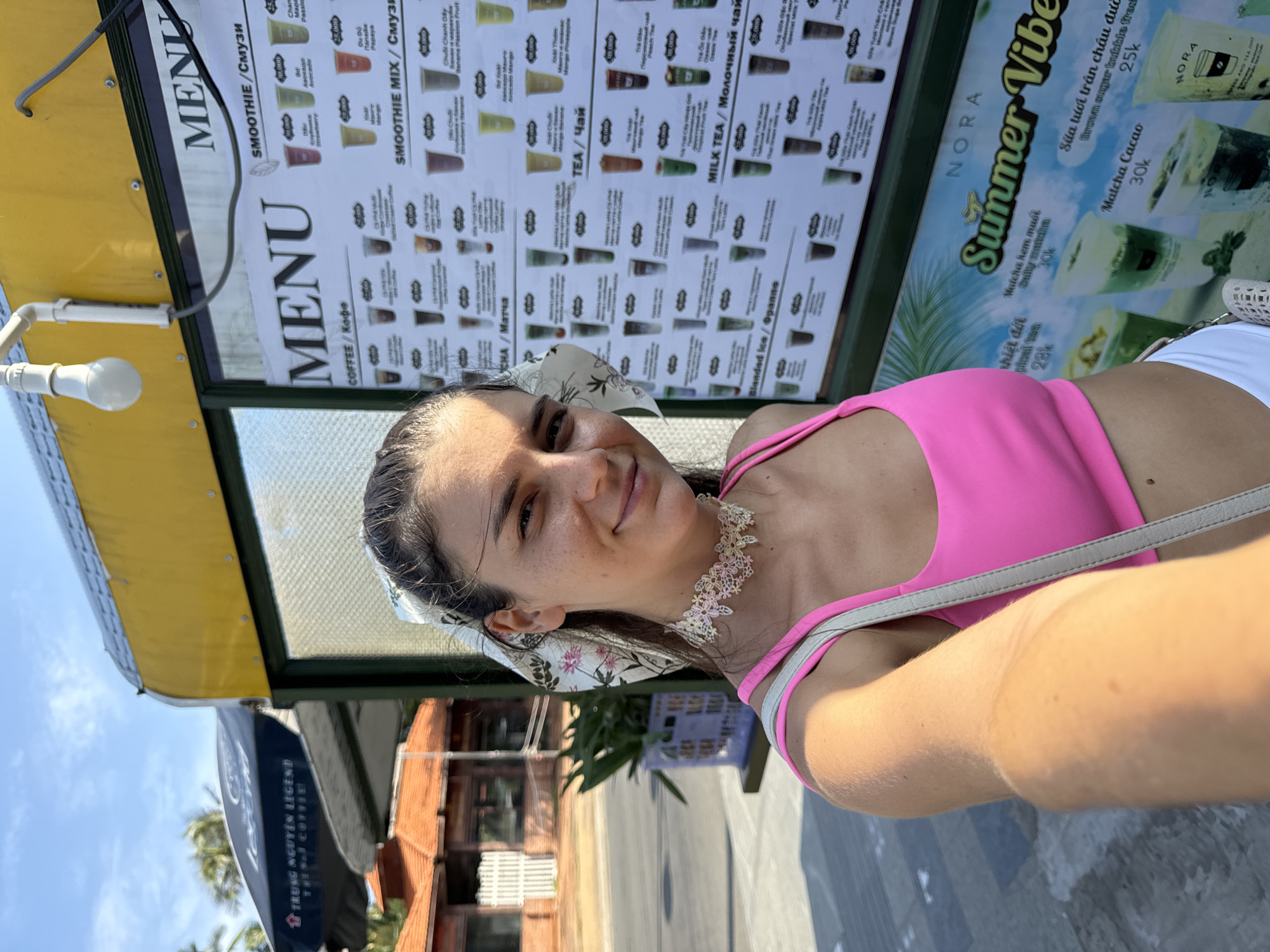 A woman in a pink tank top and white shorts takes a selfie in front of a menu board. The menu board displays various drinks, including smoothies, coffee, tea, and milk tea, with prices in Vietnamese Dong. The woman is wearing a white and pink floral headband and a white lace choker. In the background, there is a yellow awning over the menu board, a glimpse of palm trees, and buildings with orange roofs, consistent with the architecture found in Mũi Né, Vietnam. The lighting suggests it is a sunny day.