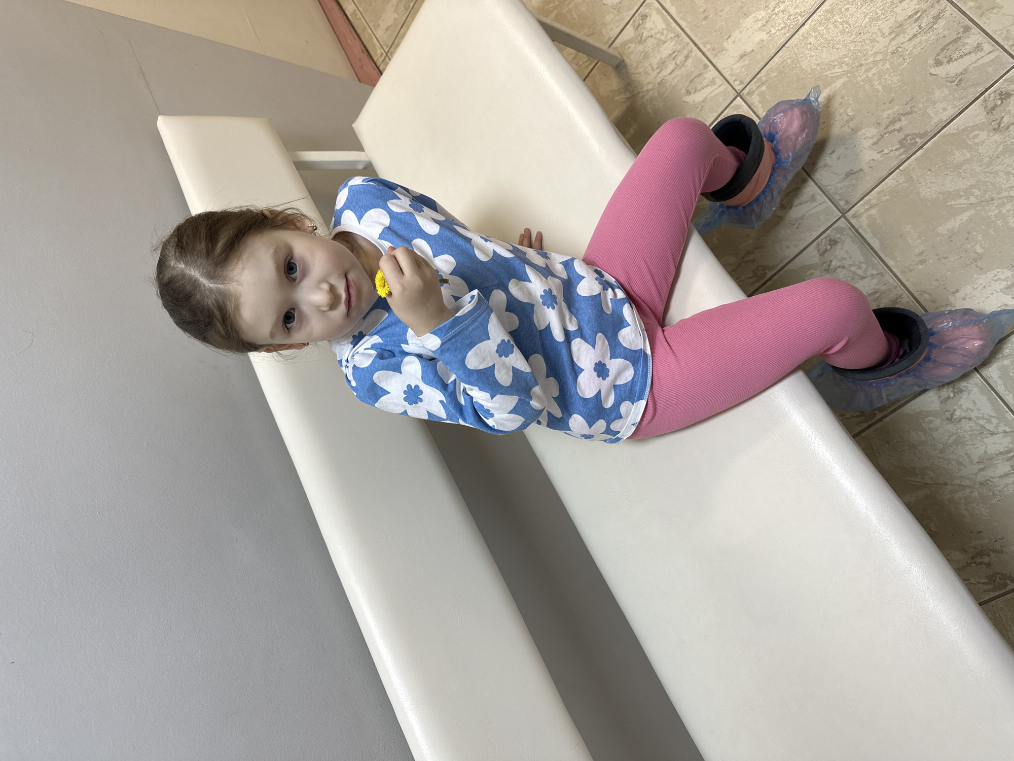 A young girl is depicted lying on a light-colored, padded bench in an indoor setting. She has dark brown hair pulled back, revealing small earrings, and blue eyes, looking directly at the camera with a calm, slightly curious expression. She is dressed in a blue long-sleeved shirt adorned with white floral patterns and bright pink leggings. Her feet are covered with blue plastic shoe covers, indicating a clean or sterile environment. In her right hand, she delicately holds a small yellow flower, likely a dandelion.
The background to the left consists of a plain light gray wall, while the floor on the right features light brown and beige square tiles with subtle veining patterns. The furniture and the presence of shoe covers strongly suggest a medical facility, such as a doctor's office or clinic, located in Chaikovsky, Russia. There is no visible text in the image, and the time of day is not discernible.