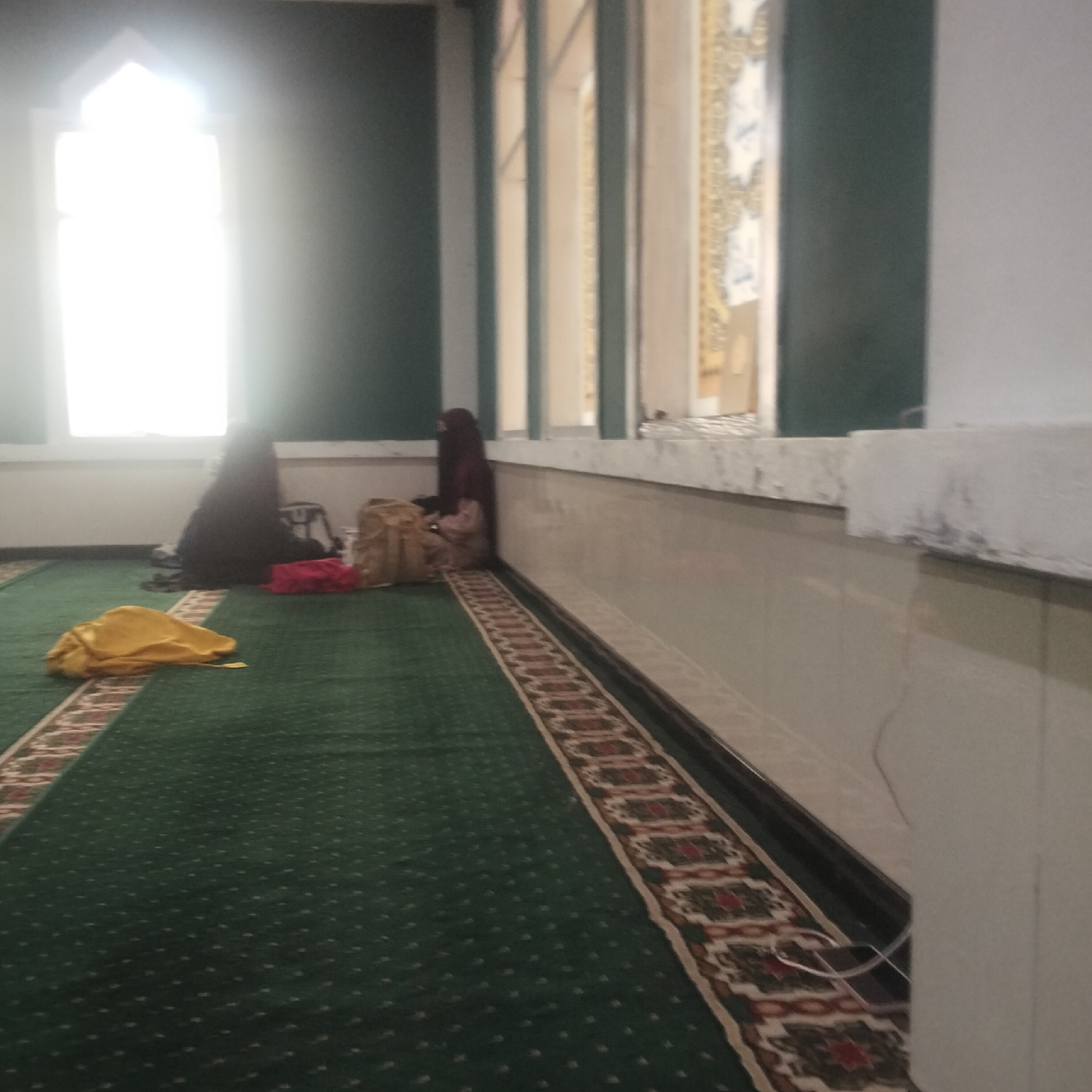 In a room with a green carpet, two women are seen sitting on the floor. One woman is wearing a dark hijab and is seated near a window. The other woman, also in a hijab, is sitting further back in the room. There are several bags and a yellow cloth on the floor near them. The room appears to be part of a mosque or similar religious building, with decorative elements visible on the walls and a patterned carpet runner. The location is identified as Cisaat, Indonesia.