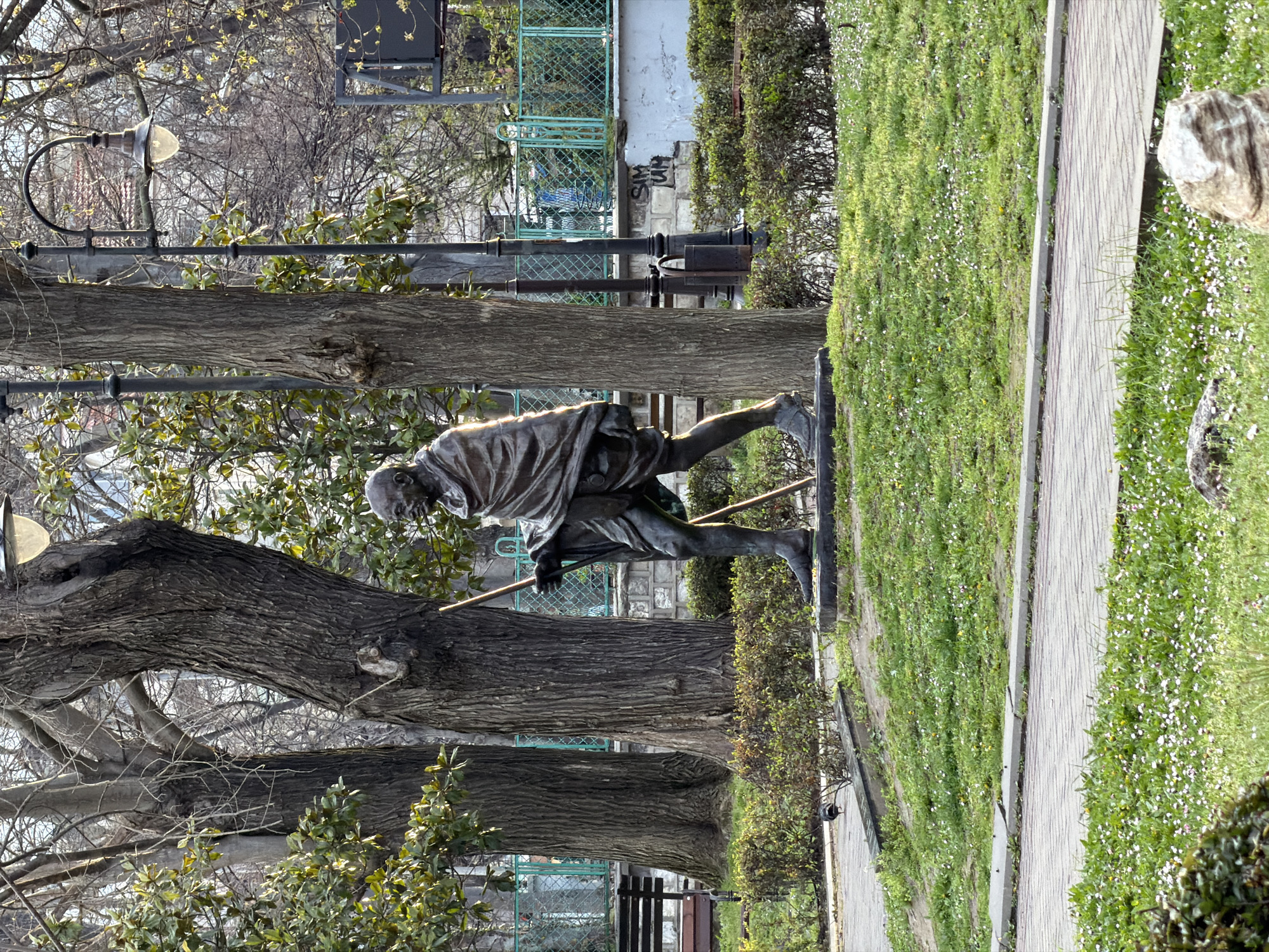 The image shows a bronze statue of Mahatma Gandhi located in a park in Varna, Bulgaria. The statue depicts Gandhi in motion, with a walking stick, wearing a traditional dhoti. The statue stands adjacent to a pathway lined with grass and small white flowers. Behind the statue are several trees with bare branches and some greenery, as well as bushes and a metal fence. In the background, there is a building or structure. The lighting suggests it is daytime.