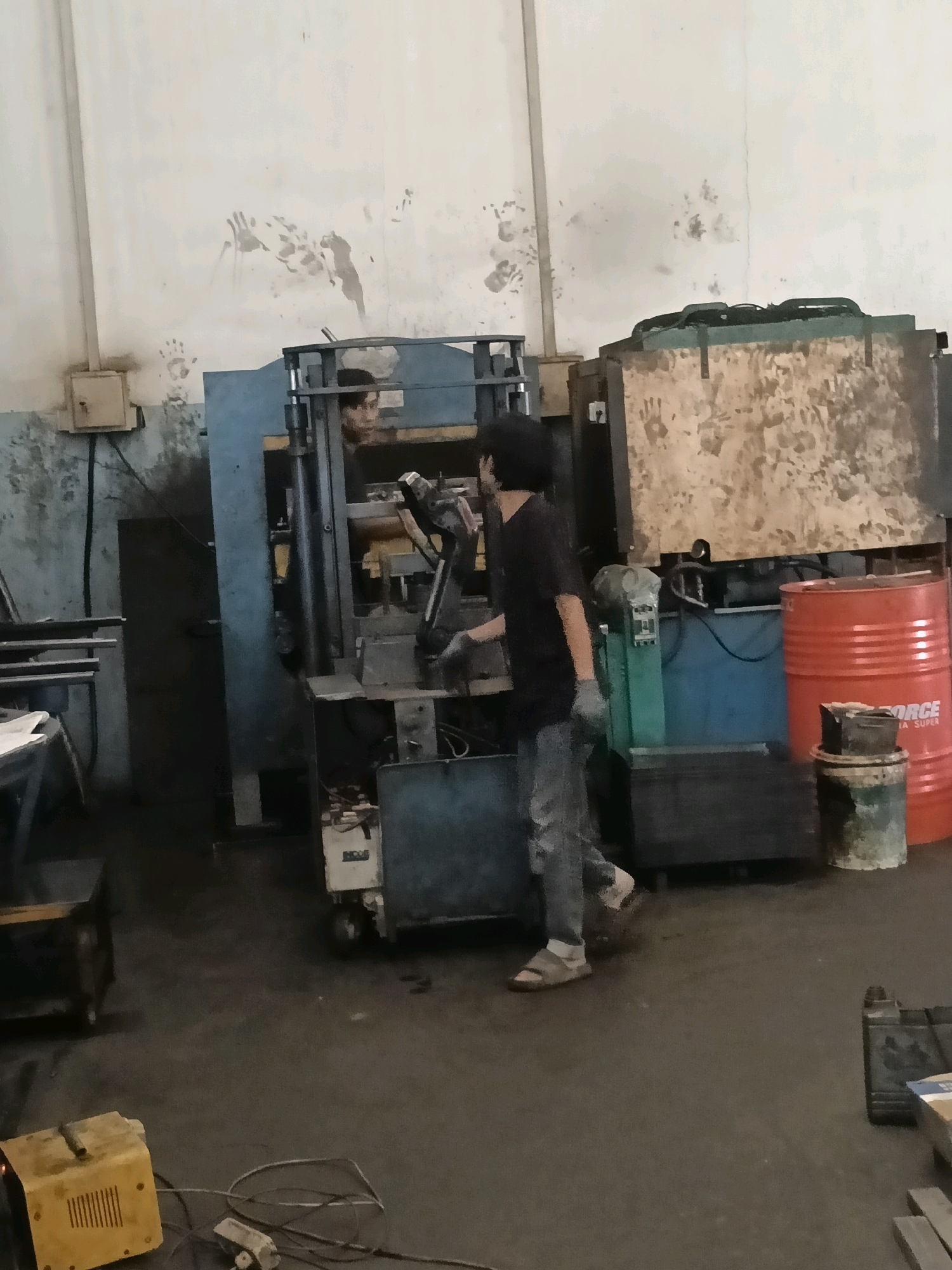 An indoor industrial setting, likely a workshop or factory, is depicted in the image. The environment appears somewhat cluttered with machinery and equipment. A prominent blue industrial press machine is visible, with a person, likely a worker, operating it or standing nearby. The worker is wearing casual clothing, including ripped jeans and sandals, and appears to be holding something in their hands. The walls of the workshop are light-colored and show signs of dirt and possibly handprints, suggesting a working environment.
In the background, there are large wooden crates or panels, some of which also have handprints. To the right, a large orange barrel, possibly containing liquid, is situated next to a smaller drum or container. The floor is concrete and appears dusty. In the foreground, a yellow electrical device, possibly a generator or power supply, is connected to cables. The overall impression is of an active, though perhaps somewhat unkempt, industrial workspace. The specific location of Belimbing, Indonesia, is not visually identifiable from the image itself, as there are no distinguishing landmarks or signage.