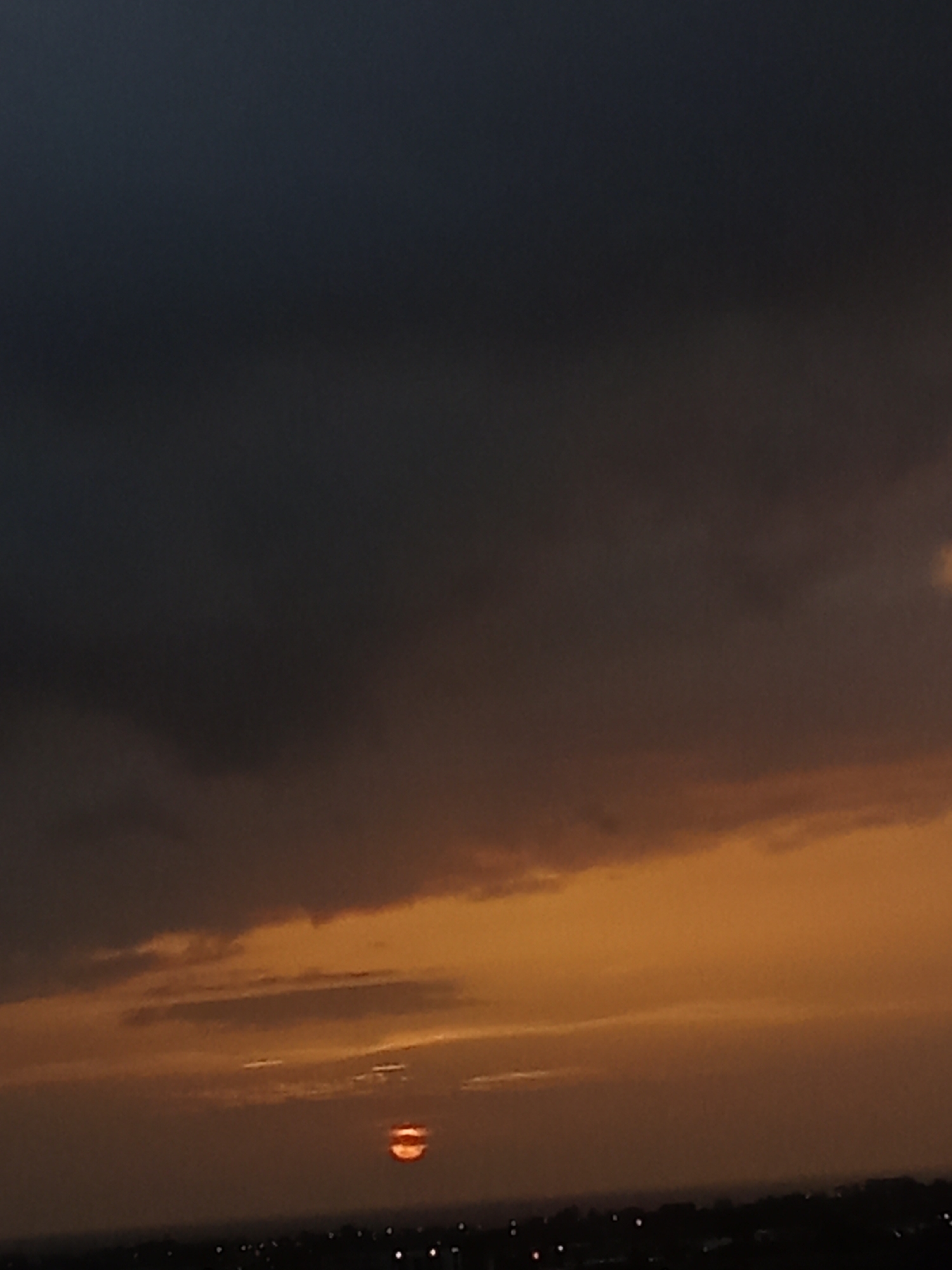 The image captures a twilight scene over a dimly lit urban landscape, presumed to be Juan Griego, Venezuela. The sky is dominated by dark, heavy clouds in the upper portion, transitioning to warmer hues of orange and yellow closer to the horizon. A small, glowing orb, likely the setting sun or a bright light source, is visible near the center, partially obscured by atmospheric haze or clouds. The foreground shows a dark silhouette of buildings and lights, suggesting the presence of a city. No people or specific objects are discernible within the image.